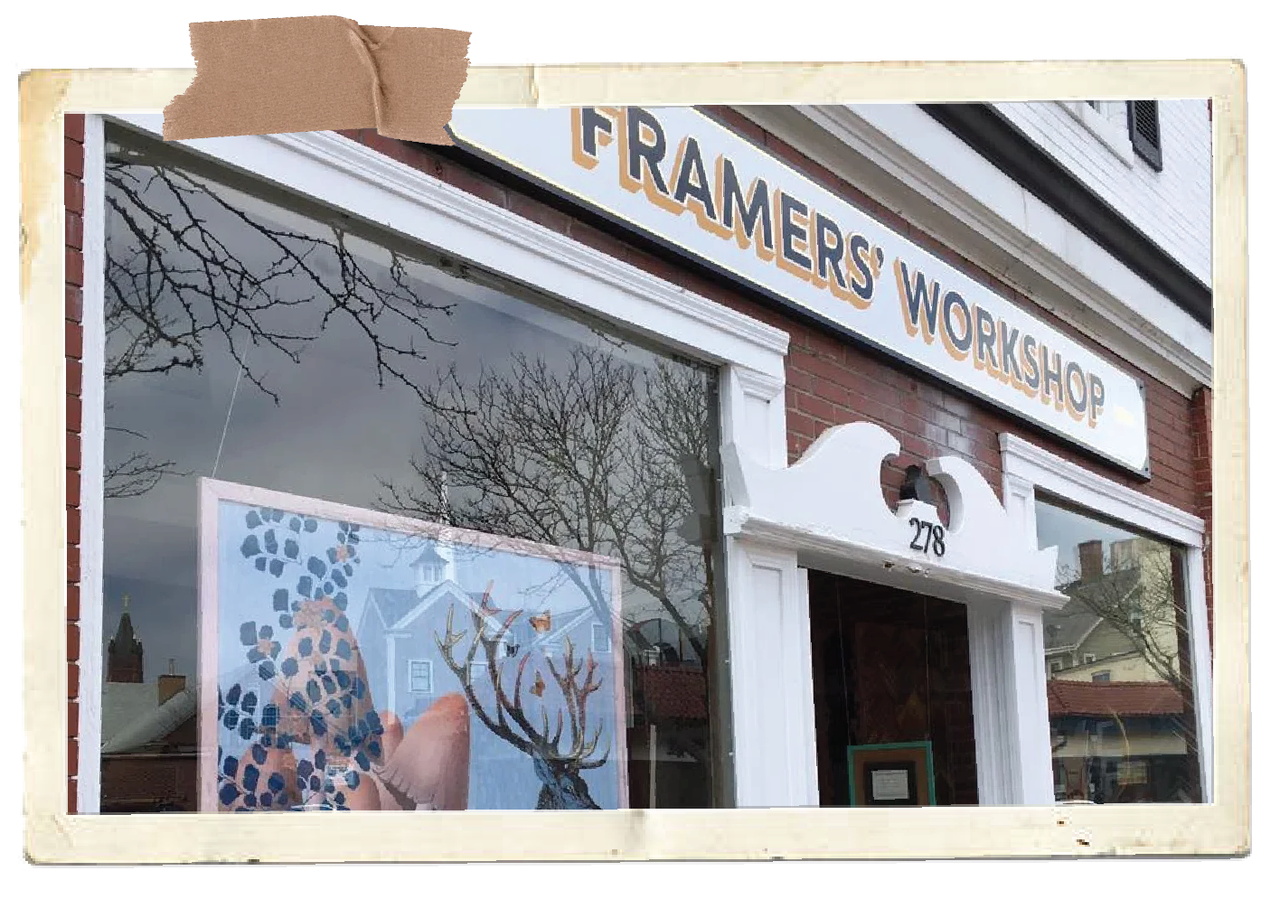 Exterior view of a storefront with a sign that reads "FRAMERS' WORKSHOP" above a large window displaying artwork, with a tree and residential buildings reflected in the glass.