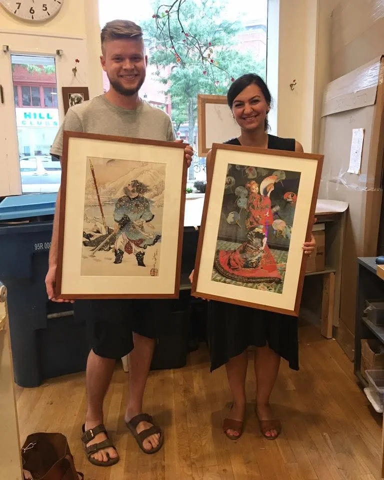 A man and woman standing inside a store, each holding framed traditional Japanese artwork. The man is on the left, wearing a light gray T-shirt, black shorts, and sandals. The woman is on the right, dressed in a black dress, open-toe sandals, and has