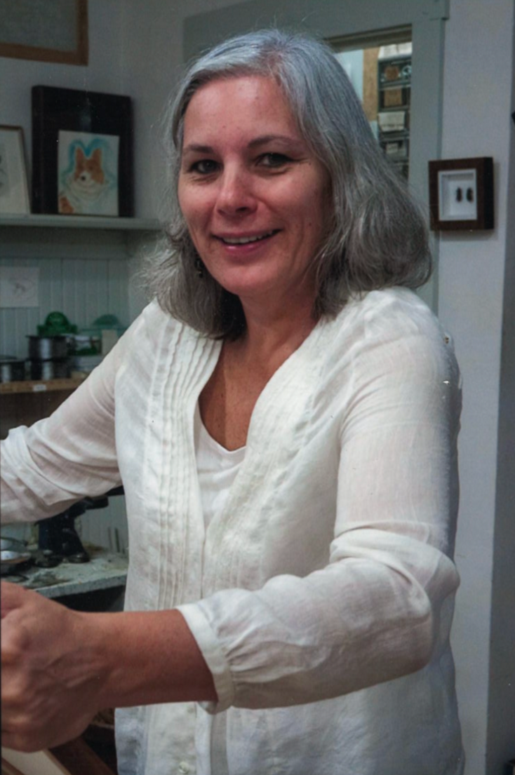 A female employee with shoulder-length gray hair smiling in a kitchen, wearing a white blouse.