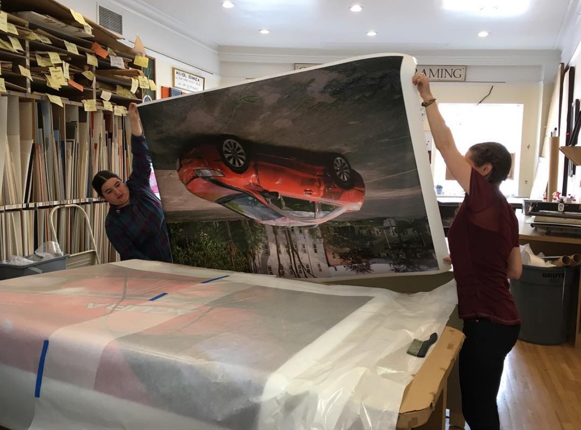 Two people are lifting and installing a large printed car photograph or poster in a workspace with shelves of rolled-up materials.