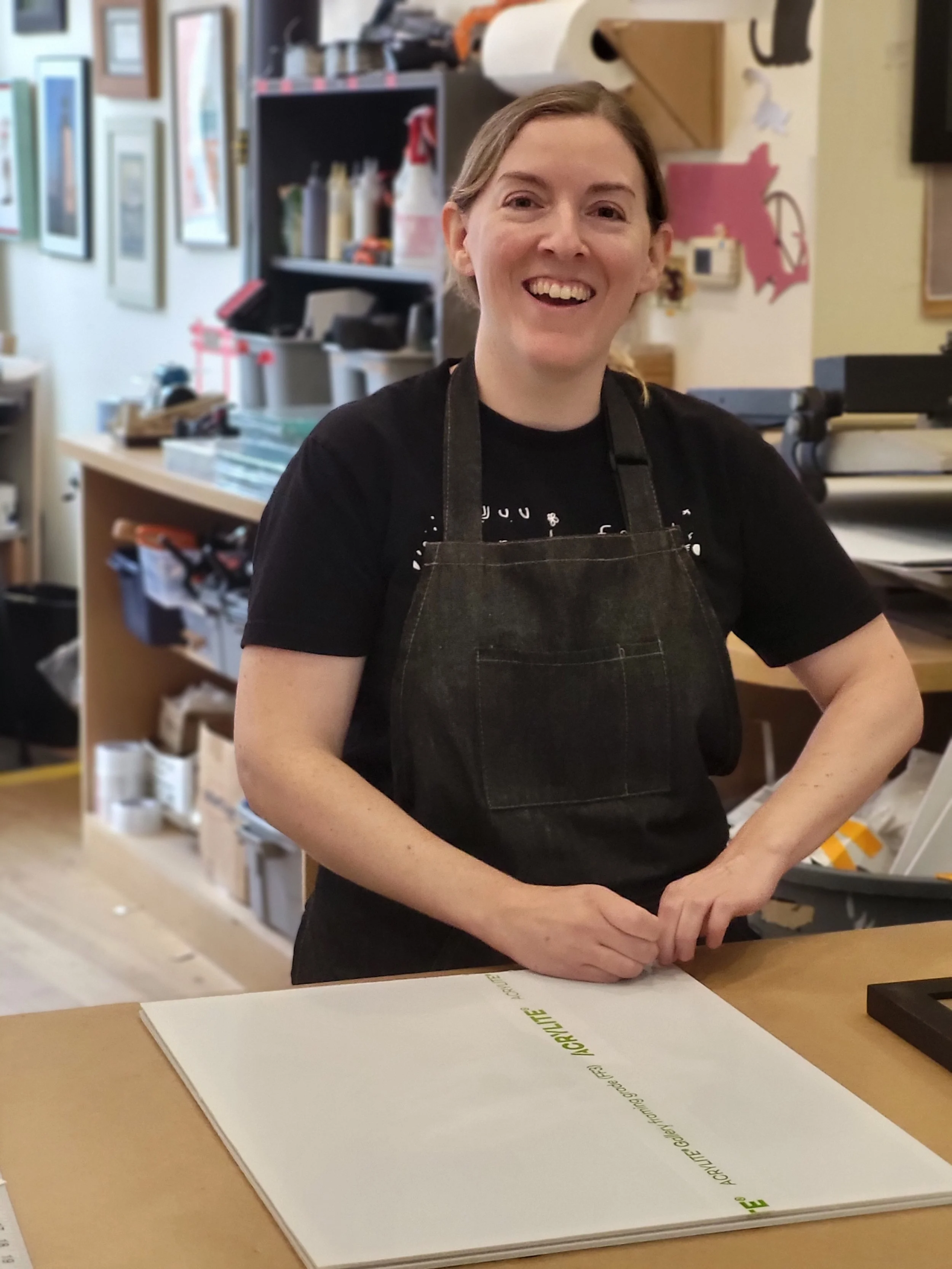 A female employee smiling in an art studio, wearing a black apron over a black t-shirt, standing at a wooden table with a large white art board.