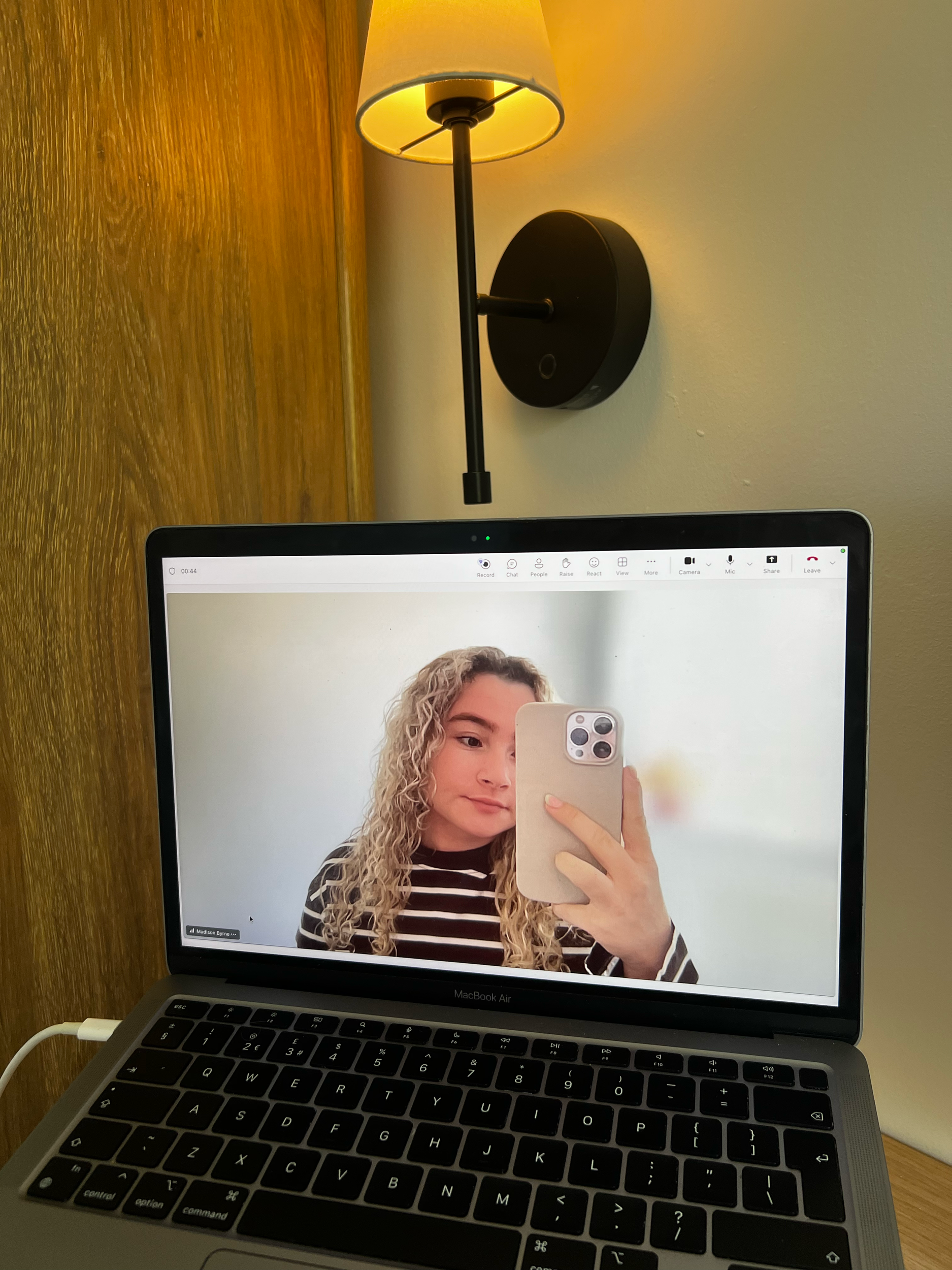 A MacBook Air laptop with an open screen showing a woman taking a selfie with her phone. The background includes a wall-mounted lamp and a wooden surface.