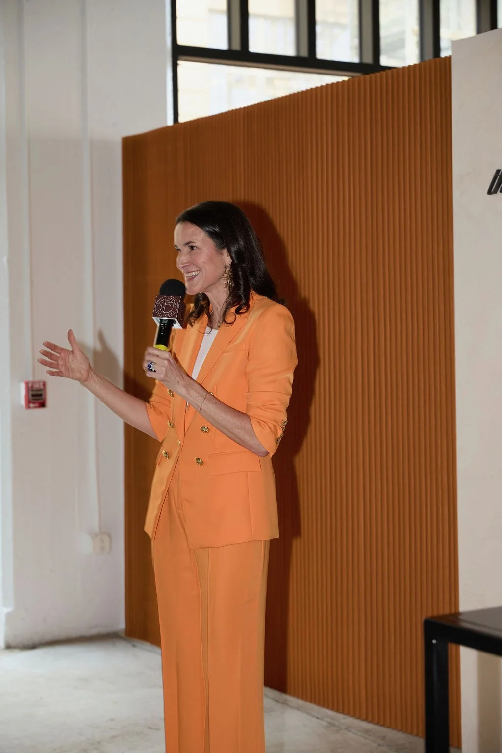 A woman with dark hair, wearing an orange suit, speaking into a microphone at an indoor event.