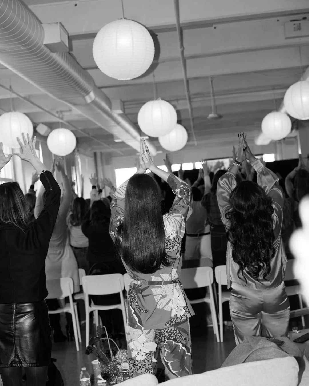 Group of people participating in a yoga or meditation class, sitting on chairs with hands raised in prayer position, in a room decorated with hanging paper lanterns.