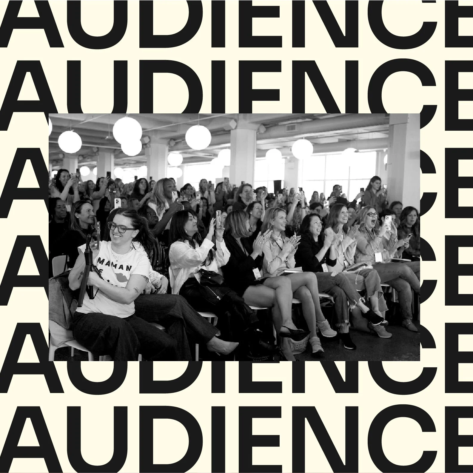 People sitting in an audience, smiling and clapping, in a bright, modern venue with hanging lights.