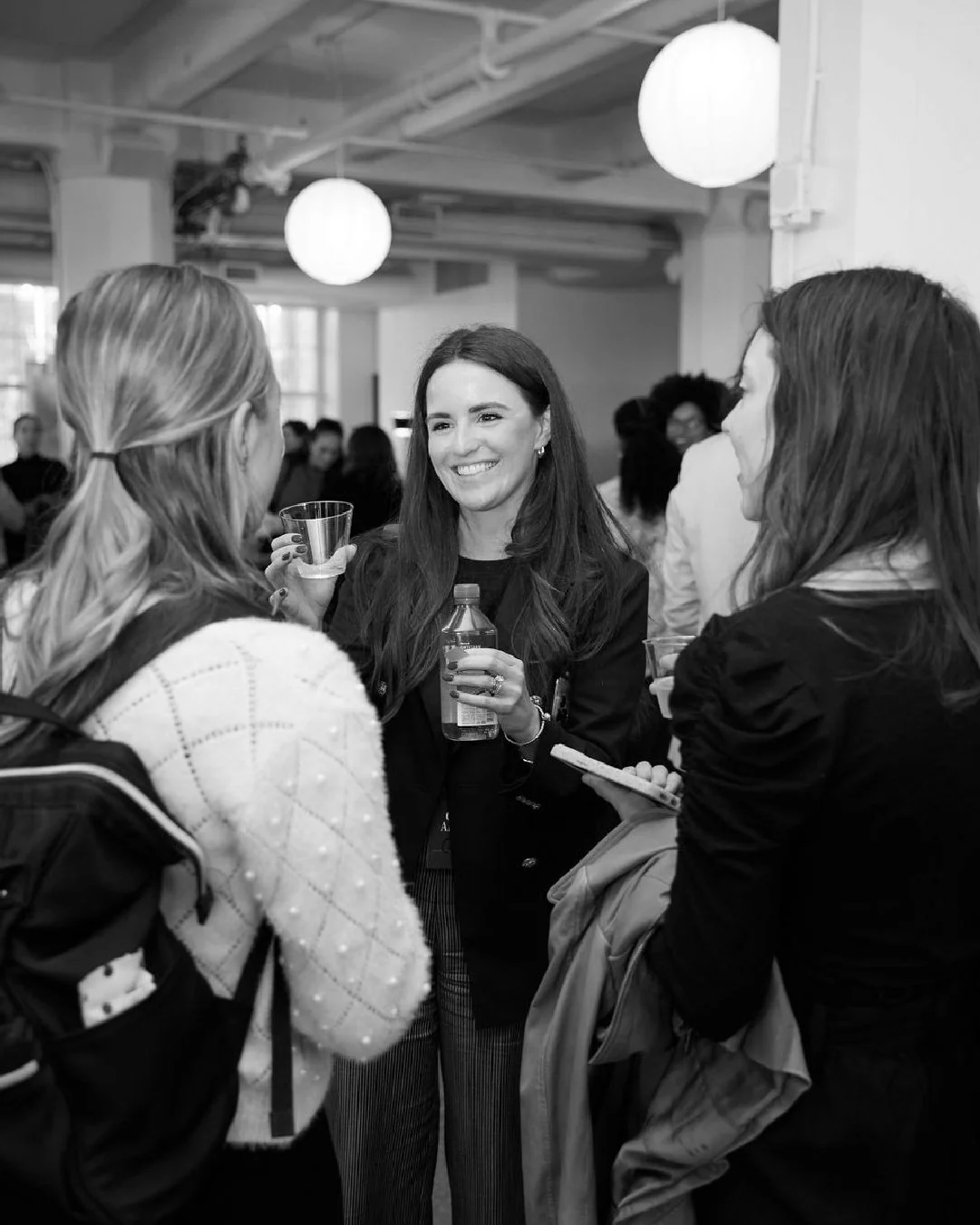 Three women having a conversation at a social event, with one smiling and holding a drink, indoors with round paper lanterns hanging from the ceiling.