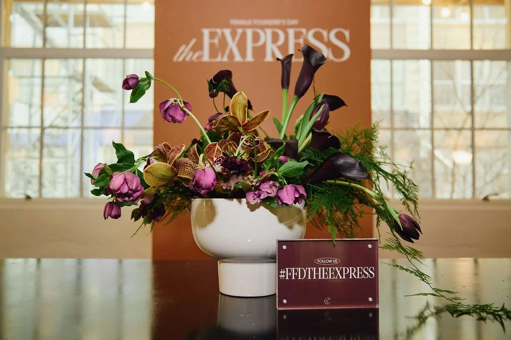 A floral arrangement with purple and green flowers in a white vase, placed on a table in front of a sign that reads '#FFD THE EXPRESS' with a blurred background of windows and natural light.