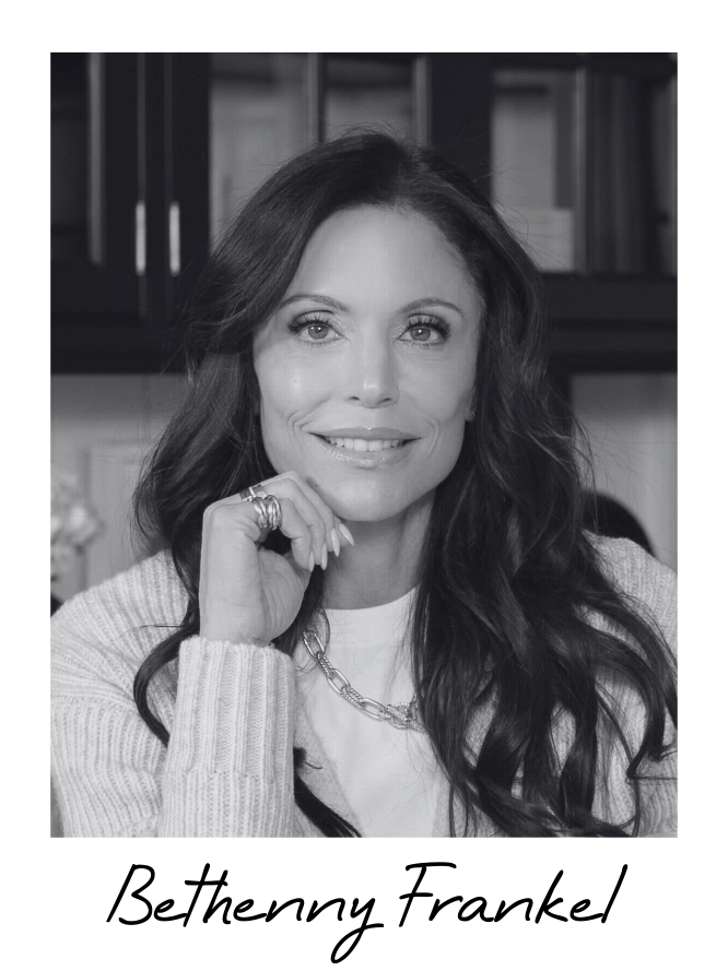A black-and-white portrait of Bethenny Frankel, a woman with long dark hair, smiling, wearing a light-colored sweater, a chain necklace, and rings, sitting in front of a bookshelf.