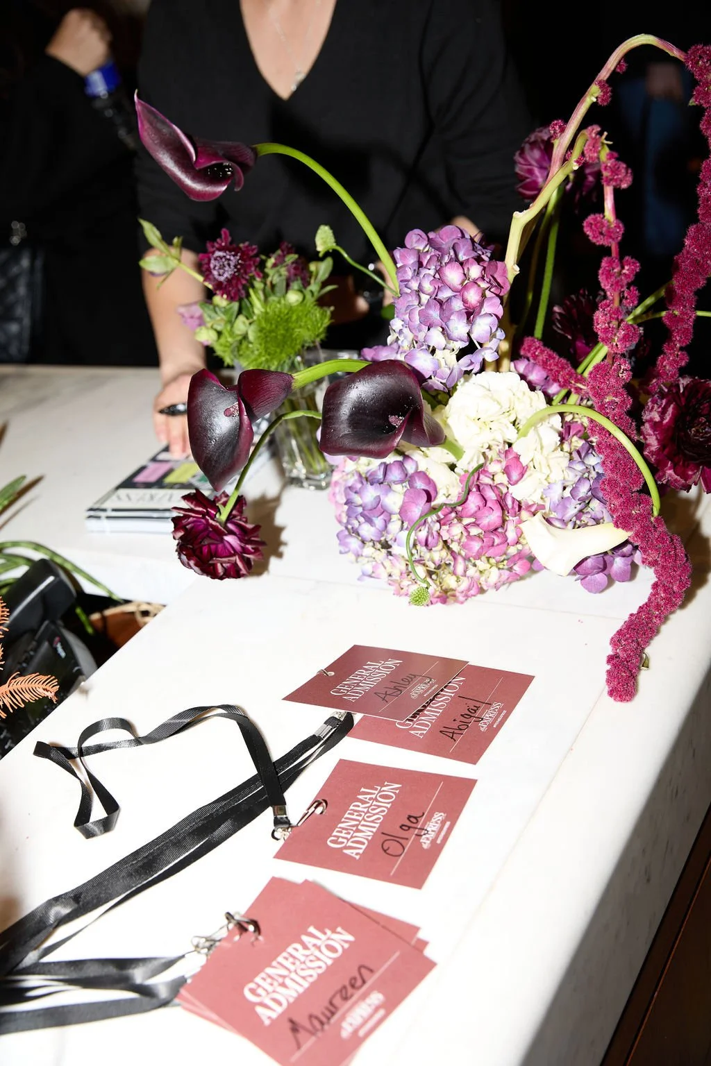 A floral arrangement with purple, white, and dark red flowers on a table with pink event passes labeled "General Admission" and "Auburn" next to black lanyards. A woman in black is behind the table, with magazines and a camera nearby.