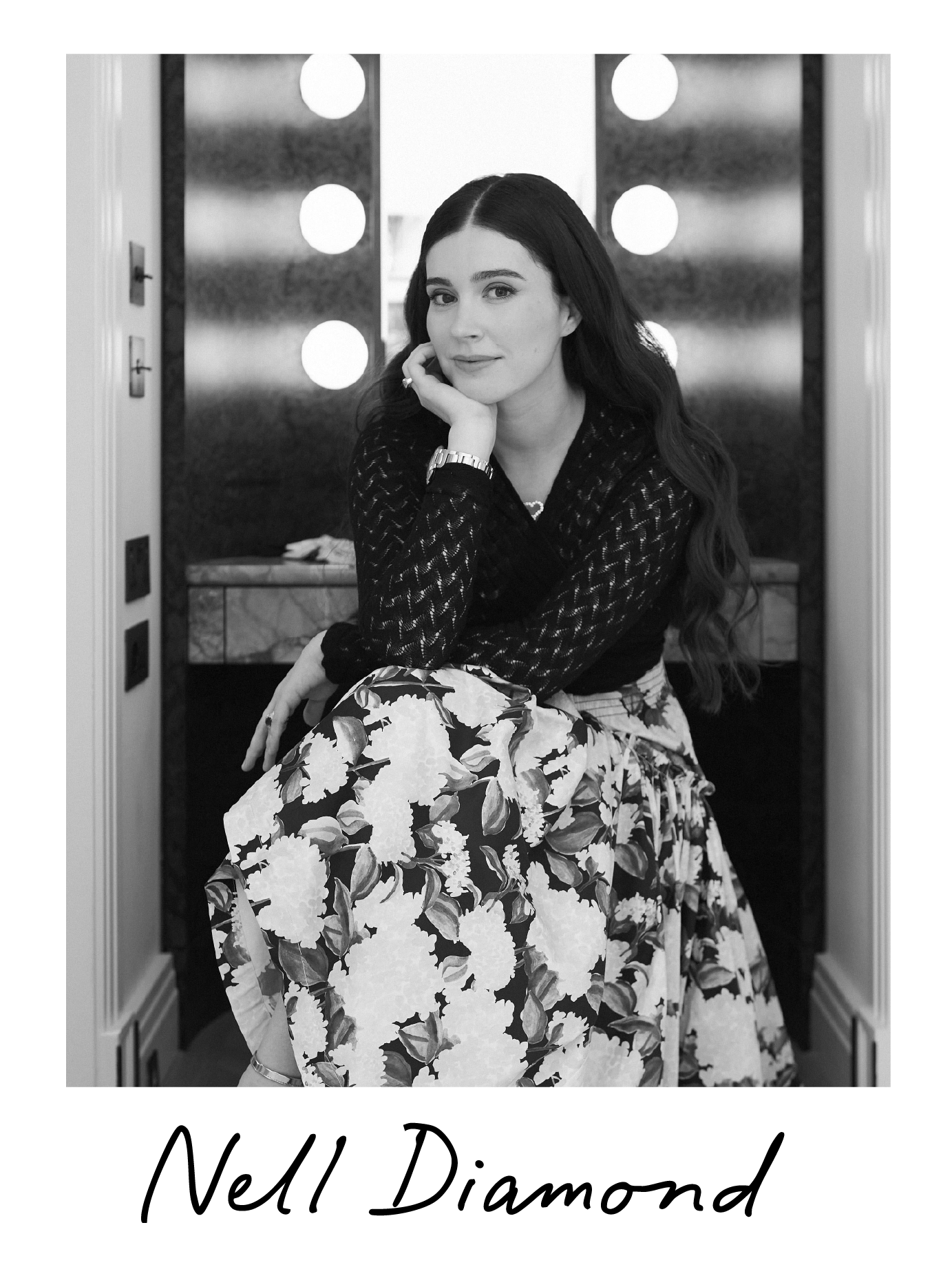A woman with long dark hair, wearing a black patterned top and a floral skirt, sitting in front of a mirror with round lights. She is resting her chin on her hand and looking at the camera. The photo is in black and white, with the words "Nell Diamond" written at the bottom.