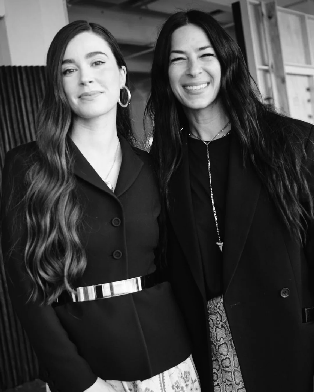 Two women standing close together, smiling at the camera; one with long wavy hair and the other with straight hair, both wearing dark clothing, in an indoor setting.
