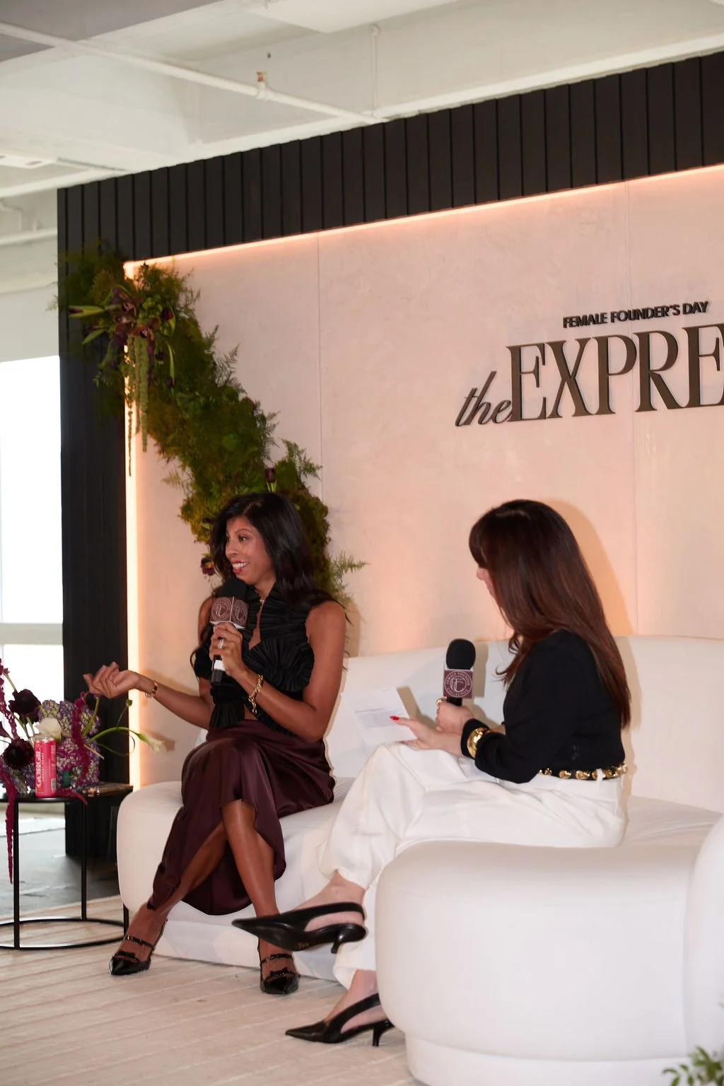 Two women are sitting on a white couch during an interview at an event celebrating Female Founders Day. One woman is speaking, holding a microphone, while the other is looking at her and holding notes. There is a floral arrangement nearby and a pink 