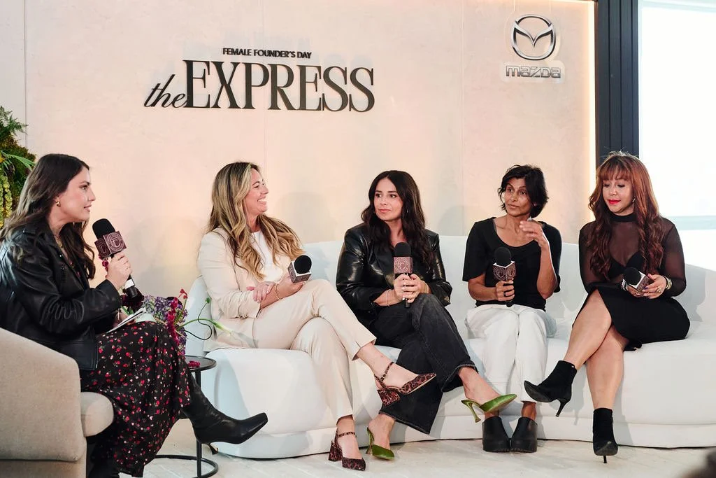 Five women seated on a white couch, holding microphones, participating in a panel discussion at Mazda Female Founders Day event. Behind them is a wall with the Mazda logo, the text 'the EXPRESS,' and 'Female Founders Day'.