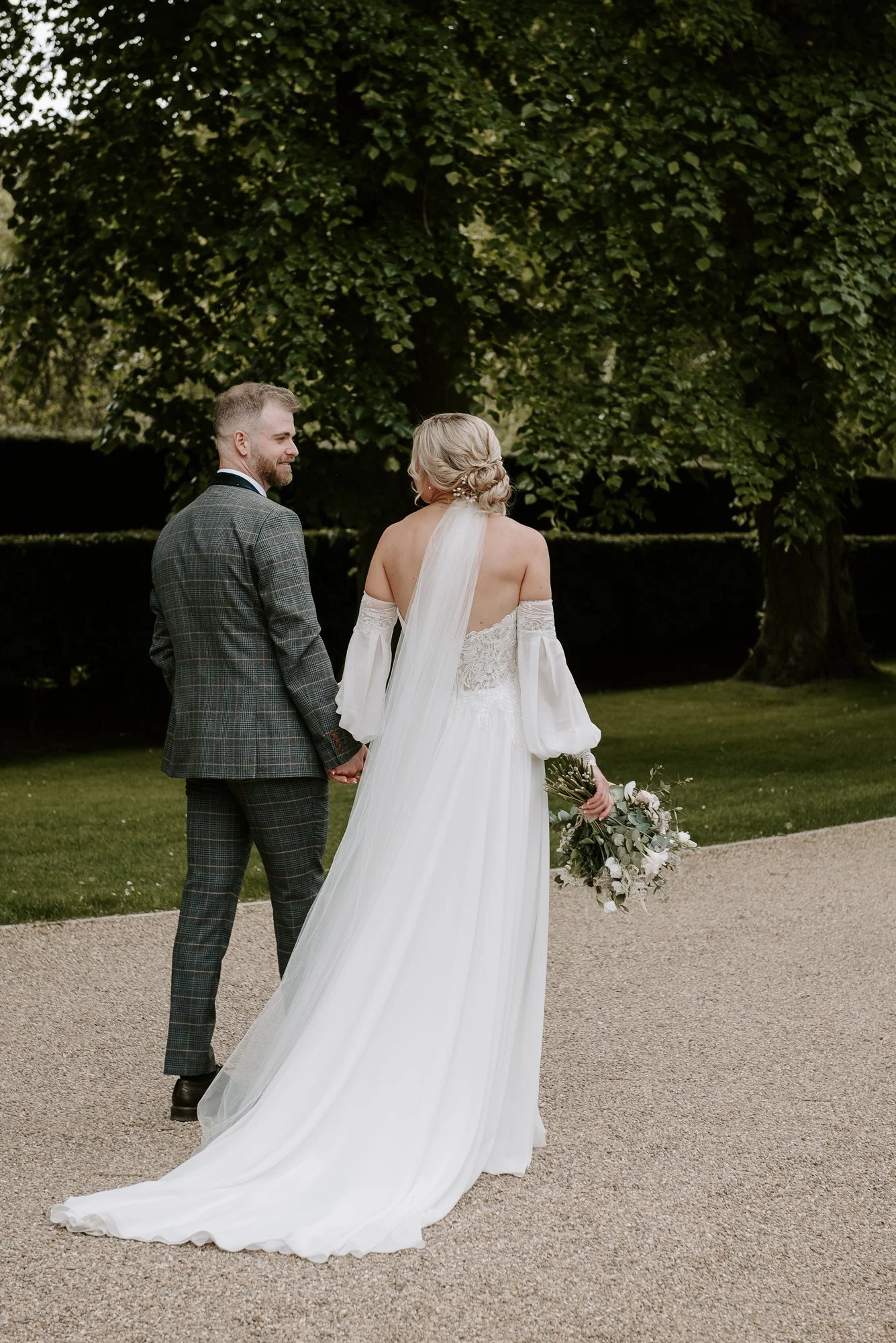 Bride and groom holding hands outdoors, bride in white wedding dress with off-the-shoulder sleeves and veil, groom in plaid suit, standing on a gravel path with green trees and grass in the background.