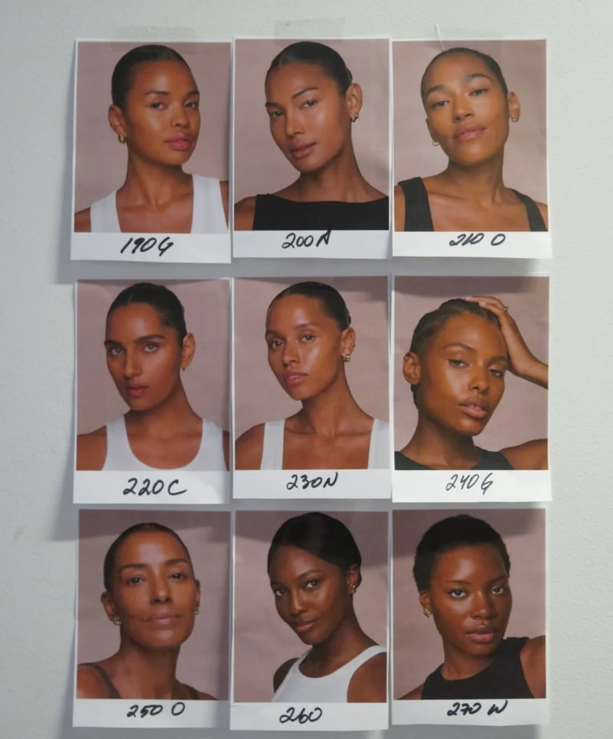 A collage of nine portrait photographs of women with different skin tones and hairstyles, each labeled with a code and some with a letter, arranged in a 3x3 grid on a wall.
