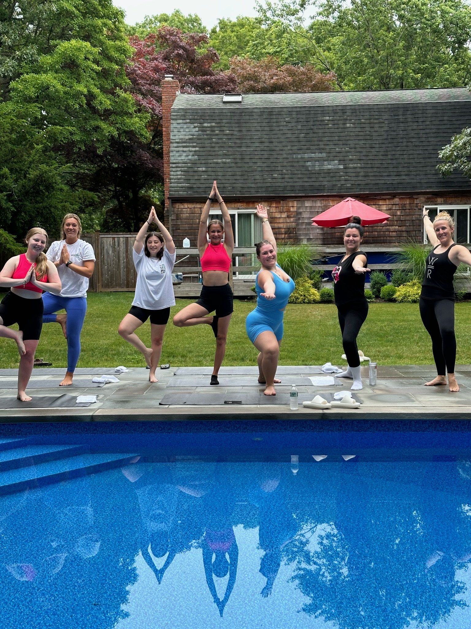 A group of seven women doing yoga or meditative poses outdoors in a backyard, with a swimming pool and lush green trees in the background.