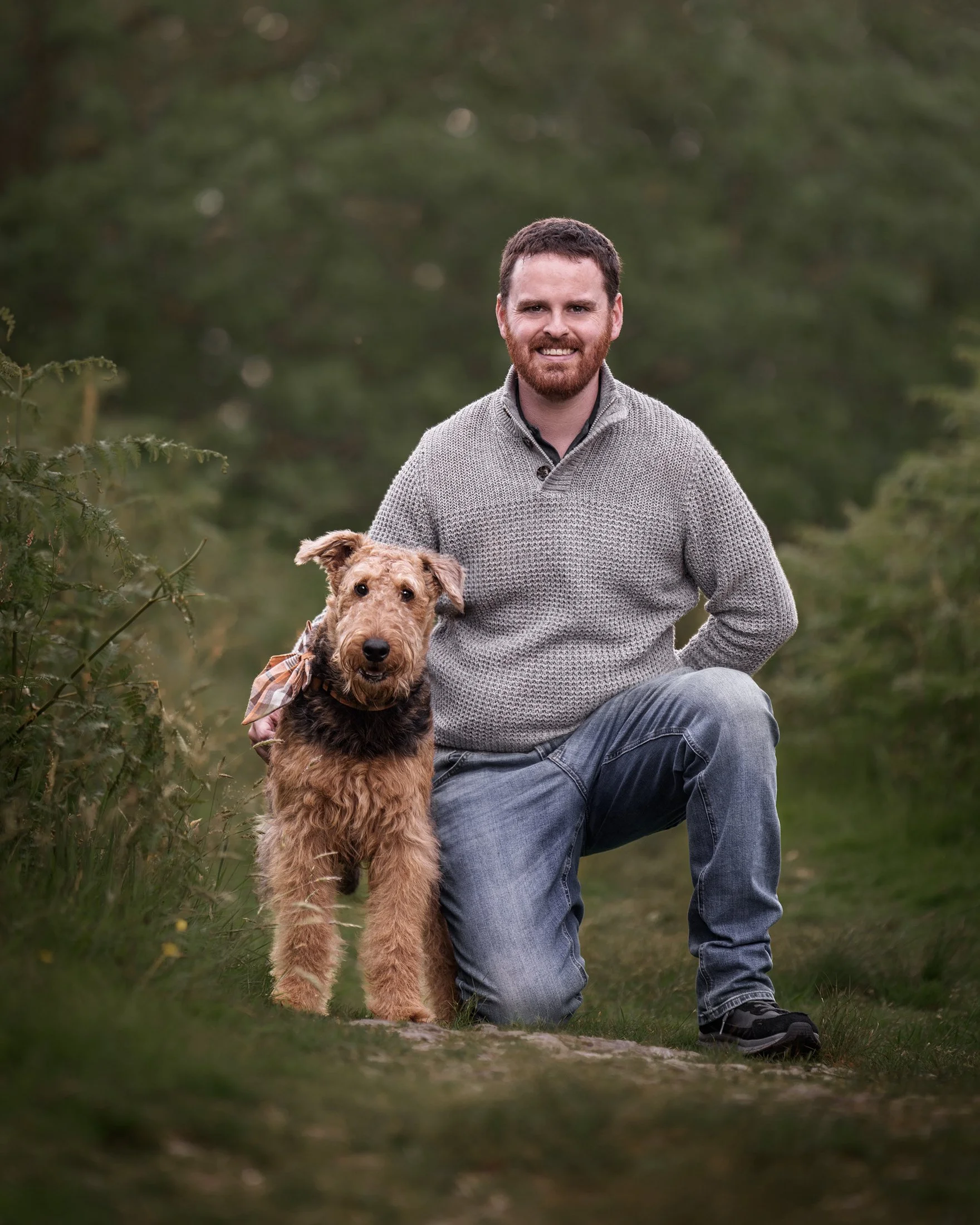 A man with dark hair, beard, wearing a gray sweater, kneeling on one knee outdoors with a dog on a walk in a green forested area.