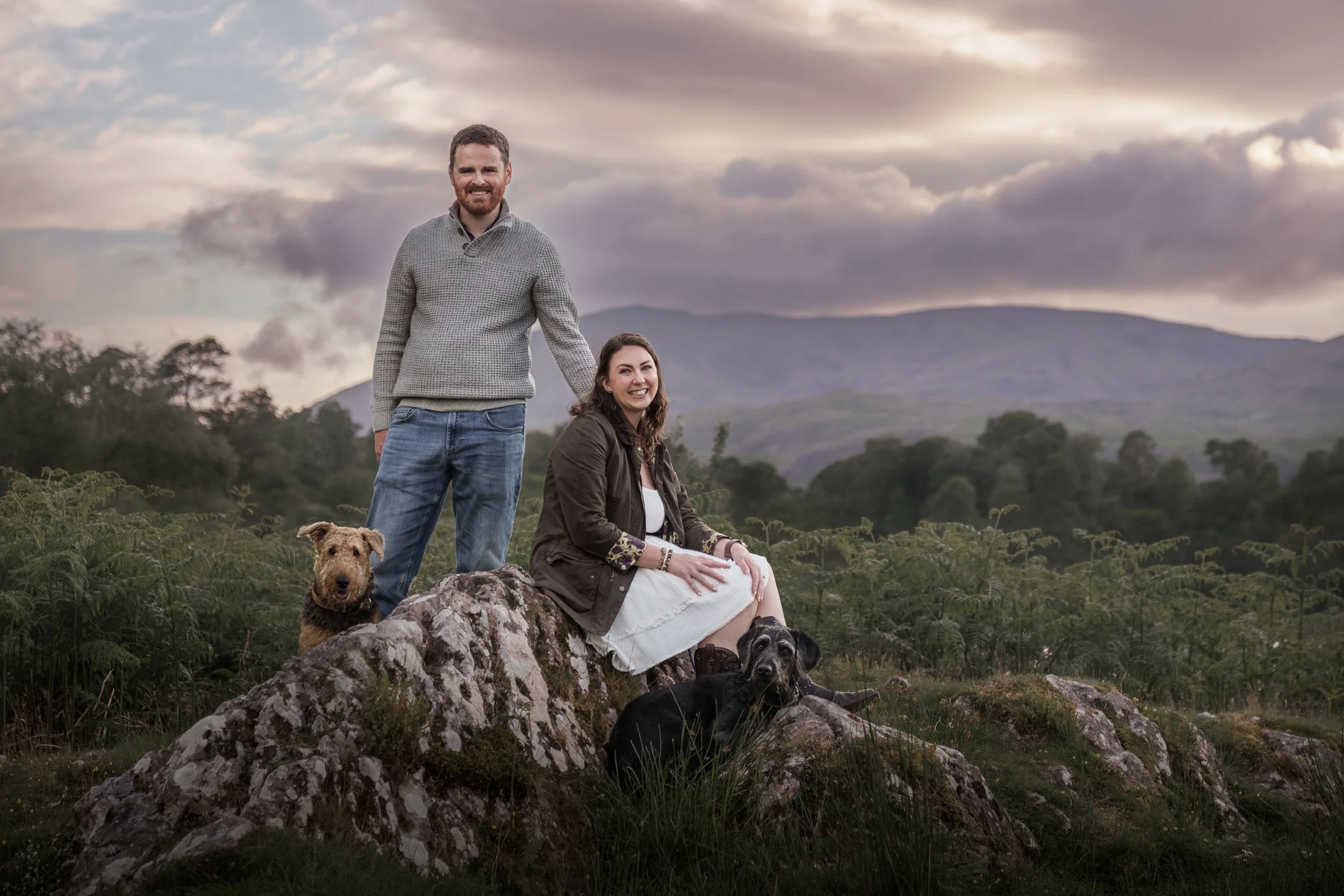 A man and woman with two dogs outdoors on rocks in a grassy field with trees and mountains in the background during cloudy weather.