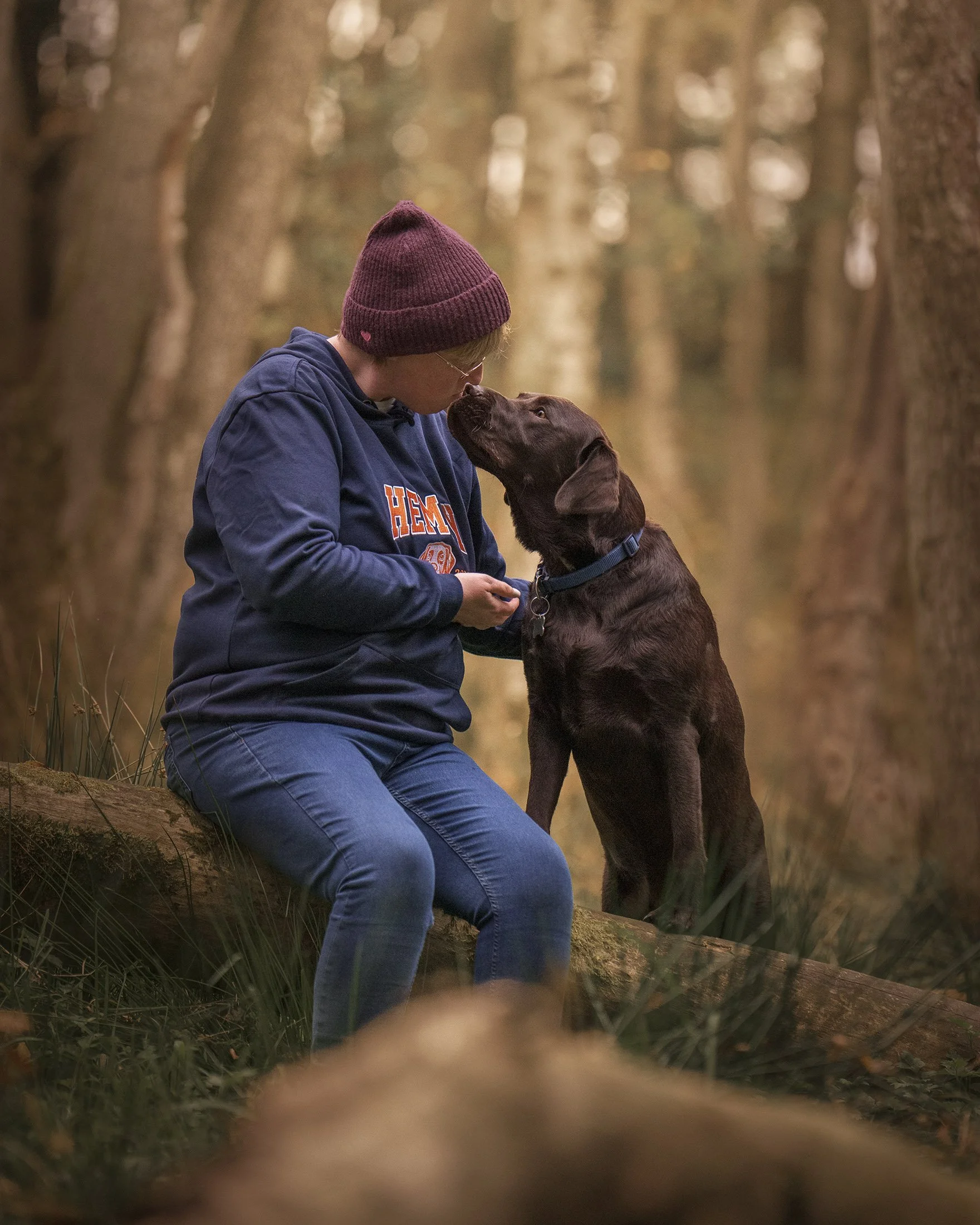 A person with a maroon knit beanie, glasses, and a navy hoodie sitting on a log in a forest, leaning in to touch noses with a brown Labrador retriever dog.