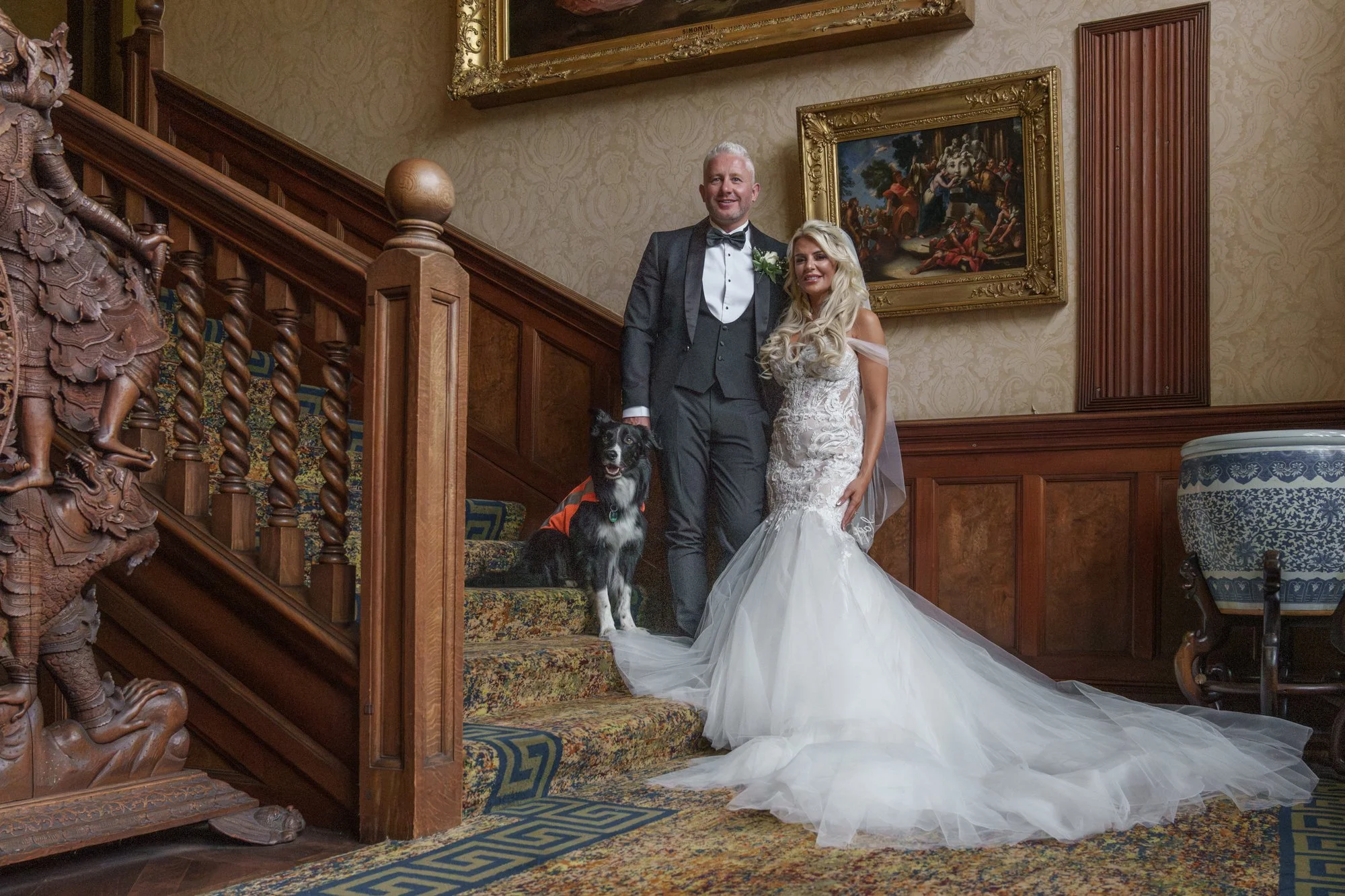 A couple dressed in wedding attire, with the man in a tuxedo and the woman in a wedding gown, standing on a staircase inside a lavishly decorated room. They are accompanied by a black and white dog sitting on the stairs. The background features ornat