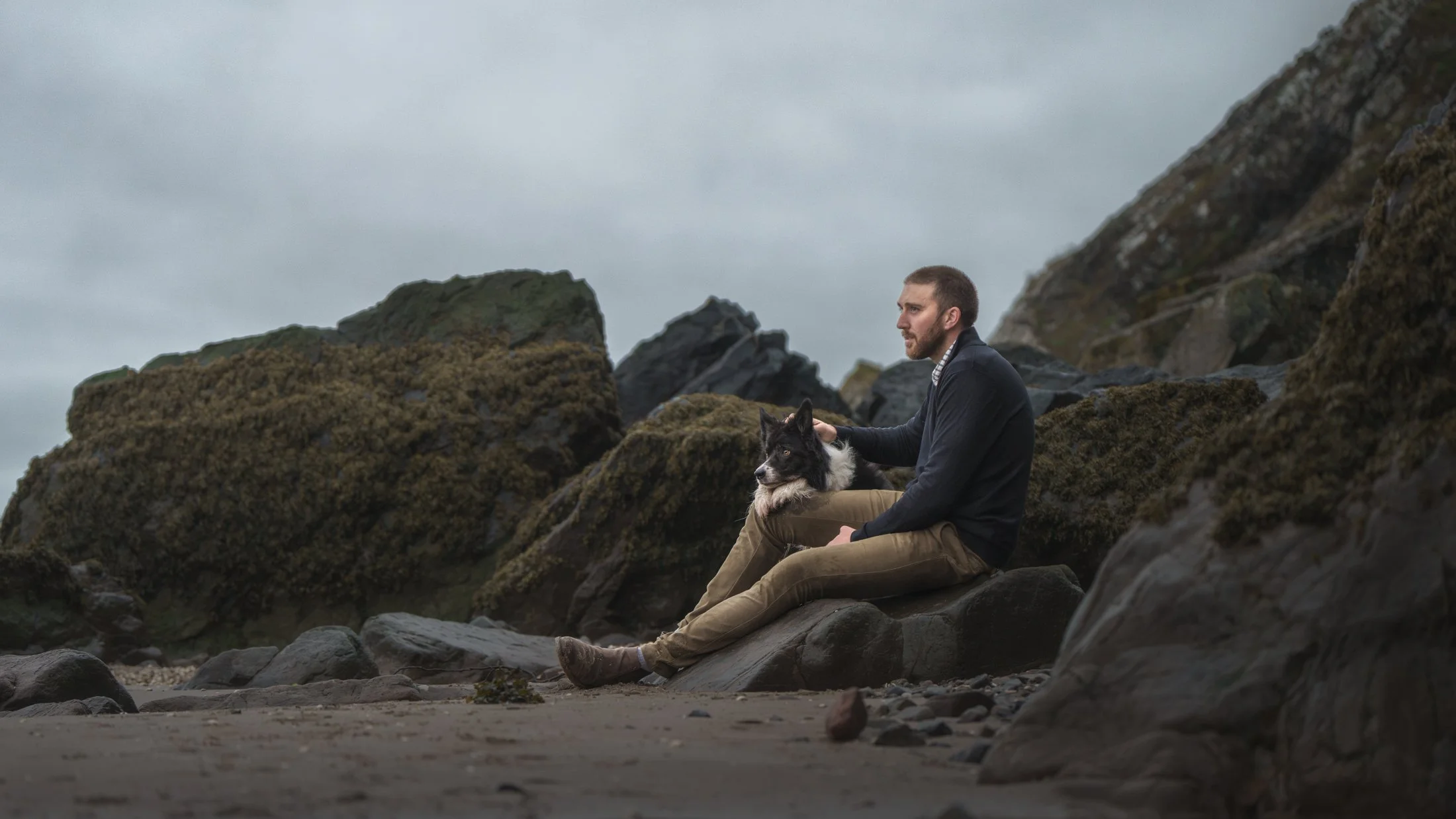A man sitting on rocks near the beach with his dog during overcast weather, surrounded by large rocks and cliffs.
