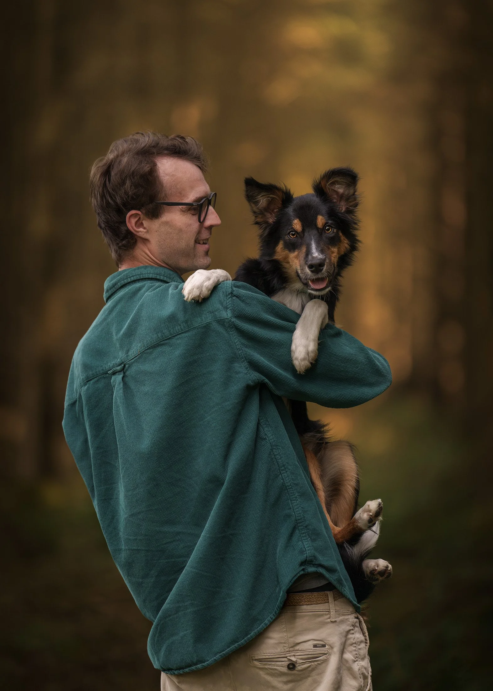 A man with glasses and casual clothing holding a dog outdoors during autumn with blurred trees in the background.