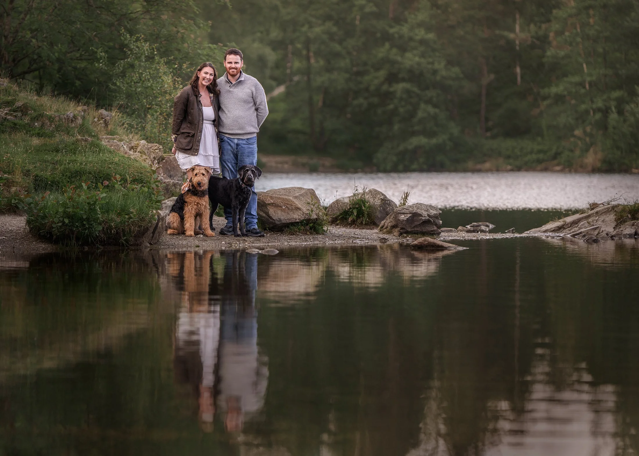 A happy couple standing by the edge of a river with two dogs, surrounded by green trees and rocks.