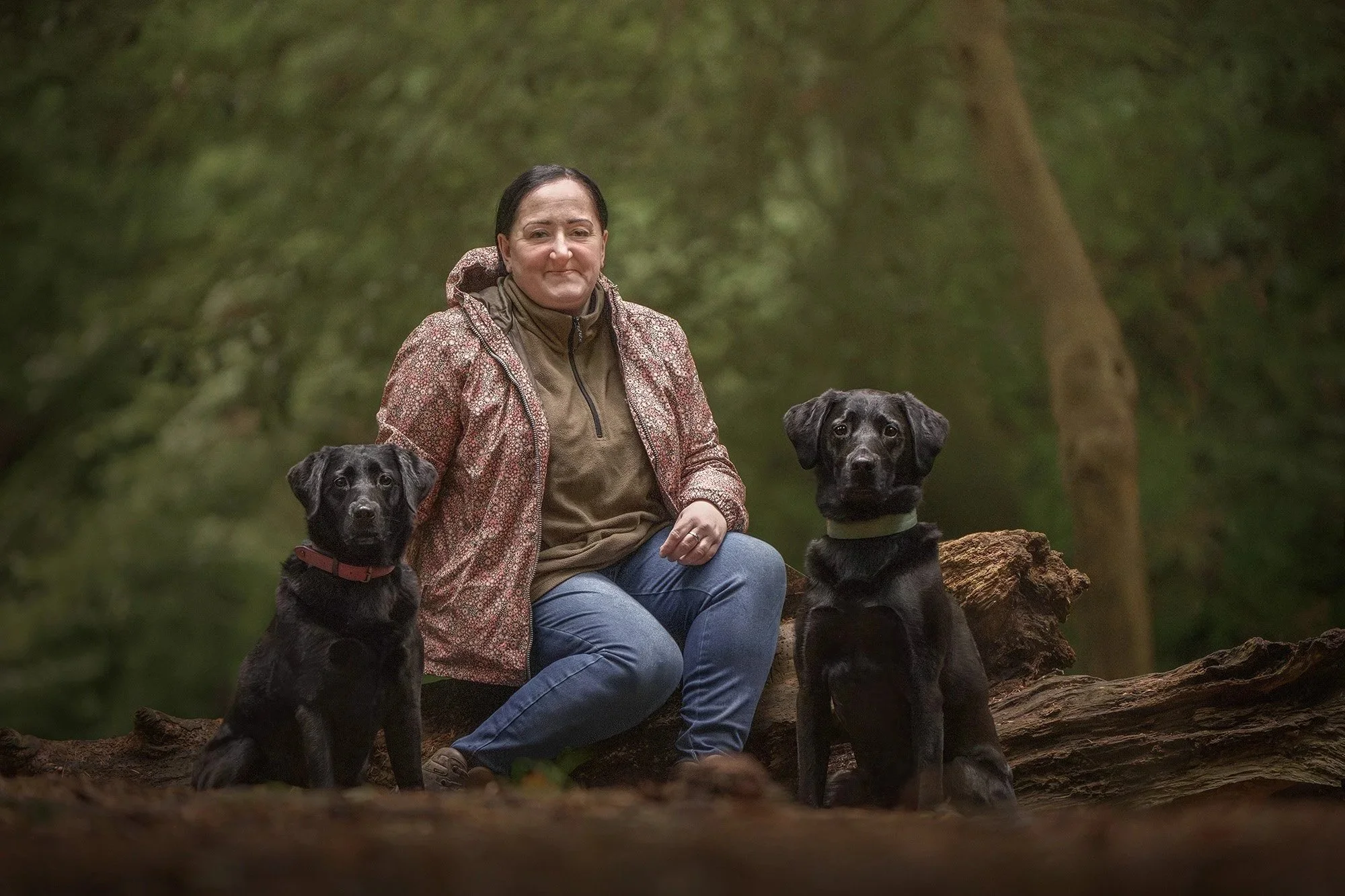 A woman with dark hair, wearing a patterned jacket over a brown hoodie and blue jeans, sitting on a log in a forest with two black dogs sitting beside her.