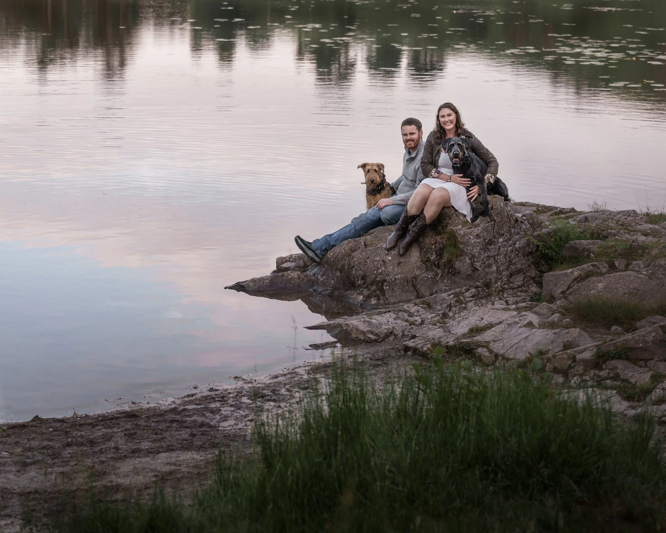 A couple with two dogs sitting on a large rock by a lake during sunset.
