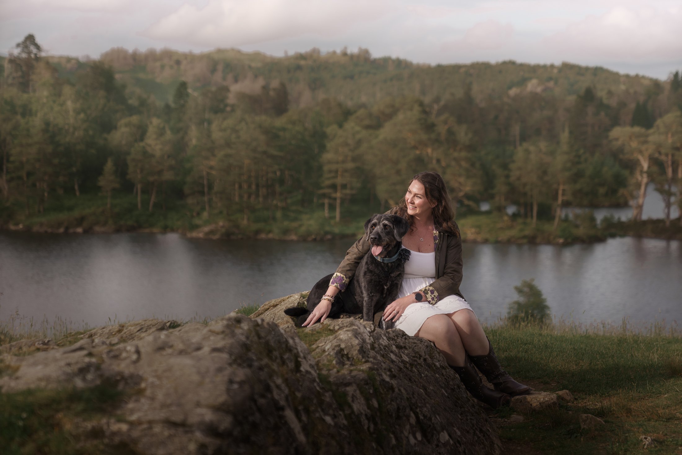 A woman sitting on a rock by a lake, holding a black dog, with a background of trees and hills under a cloudy sky.