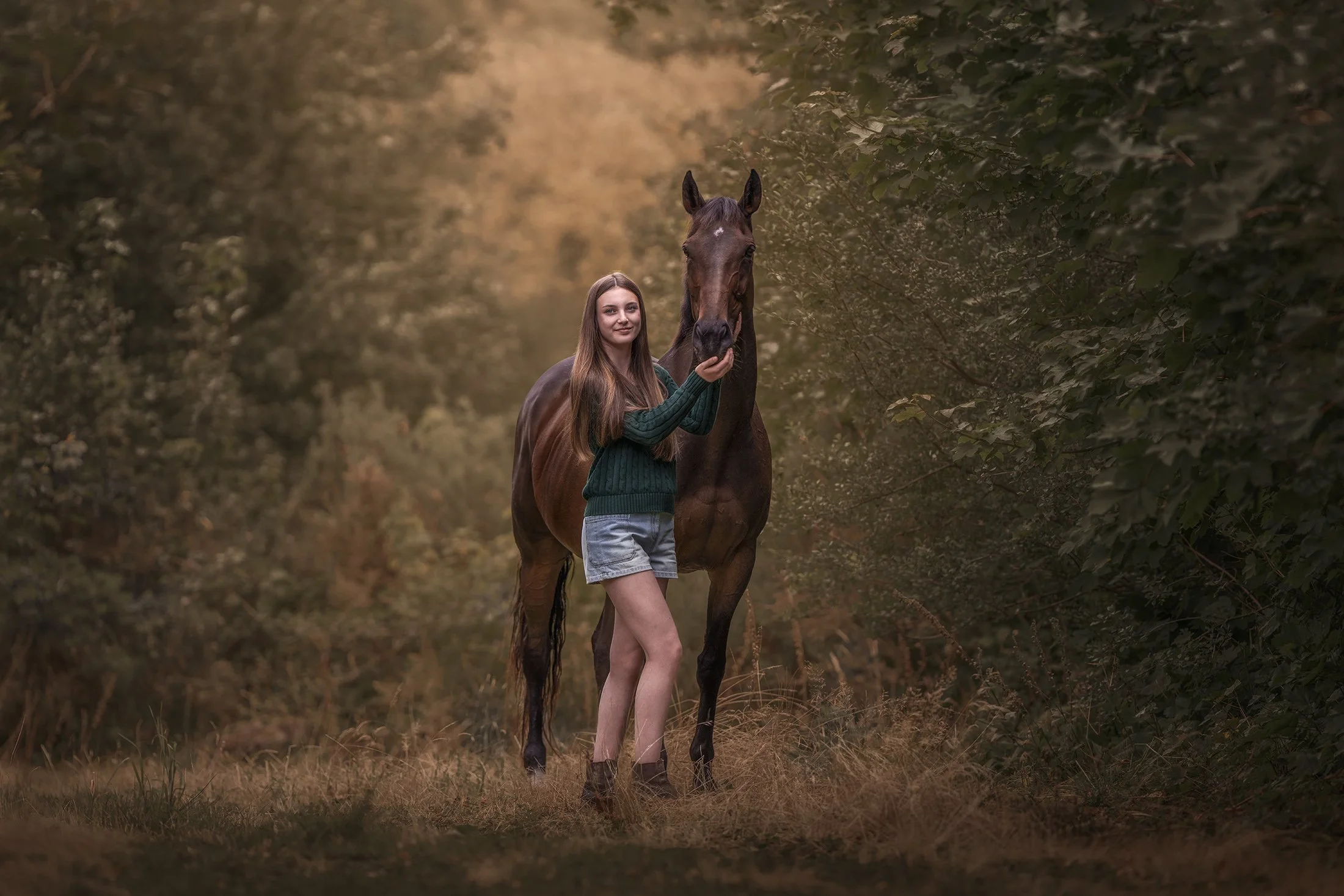 A girl with long brown hair in a green sweater and shorts standing next to a brown horse in a wooded area with trees and foliage.