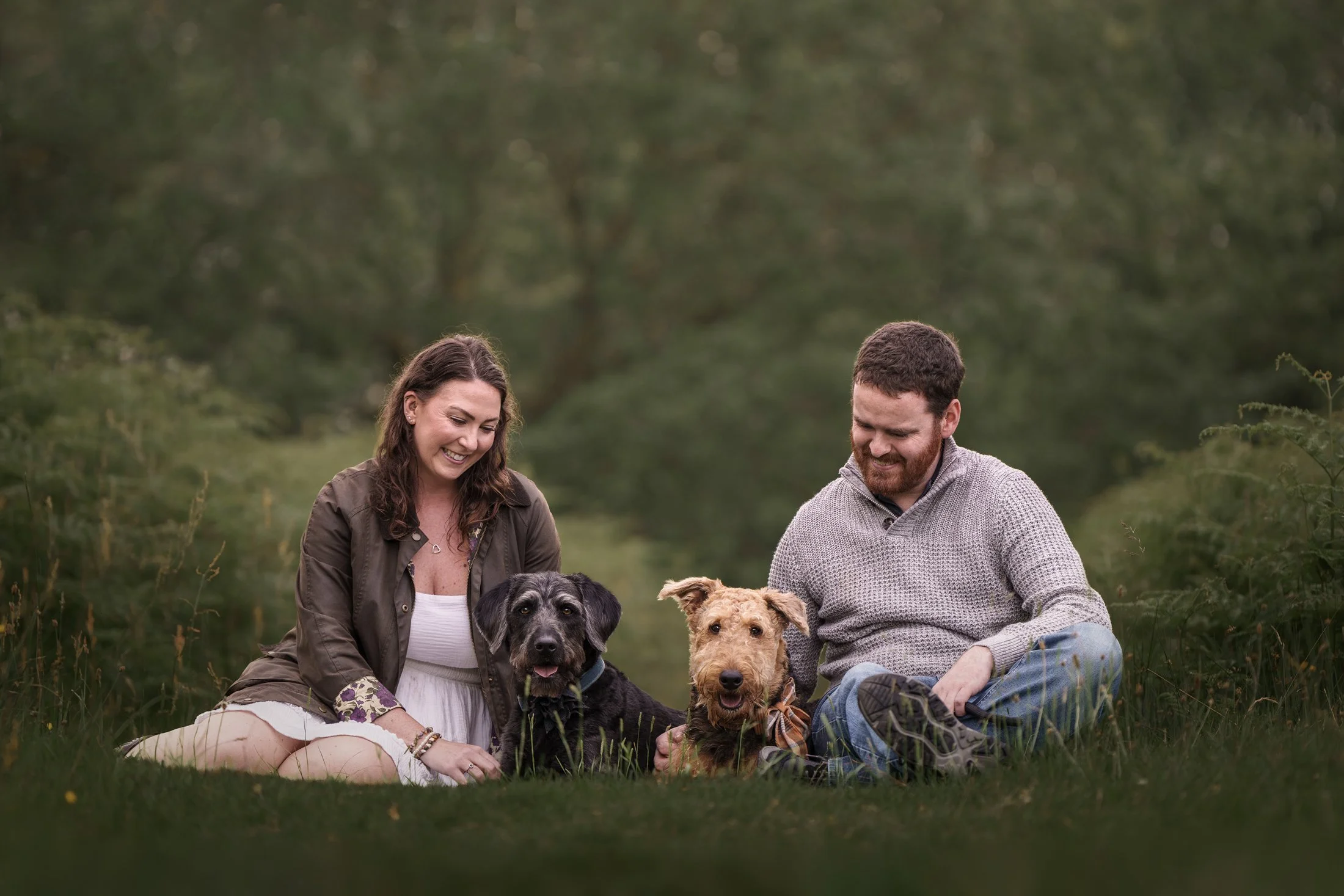 A woman and a man sitting on grass outdoors with three dogs, smiling and interacting with the dogs, surrounded by greenery.