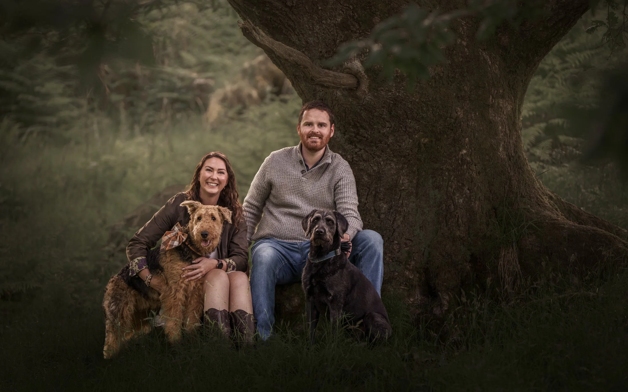 A smiling woman and a smiling man sitting outdoors under a large tree, accompanied by three dogs, one golden and two black, in a forested setting.