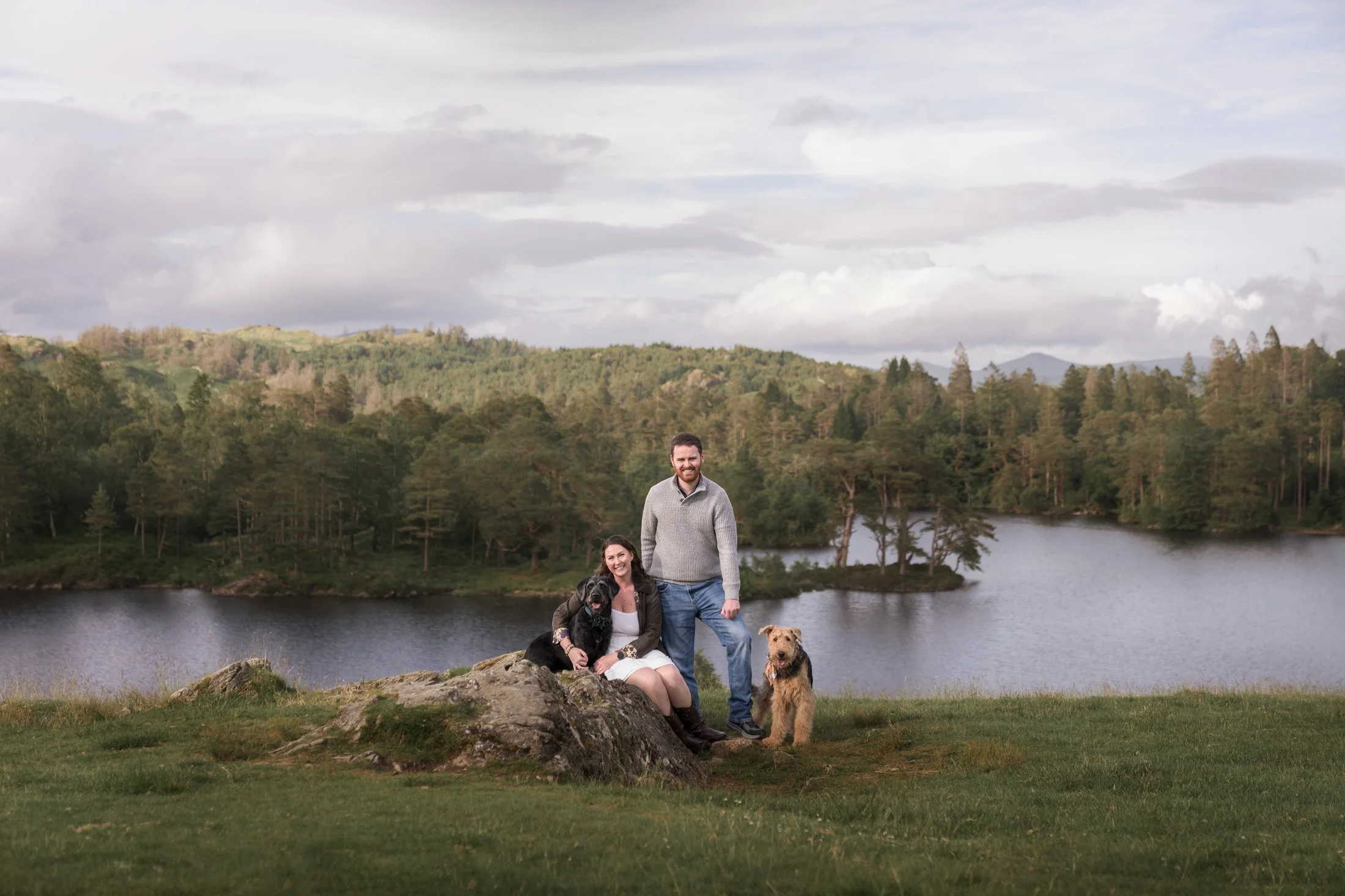 A man and woman with two dogs by a lake, with trees and hills in the background, under a partly cloudy sky.