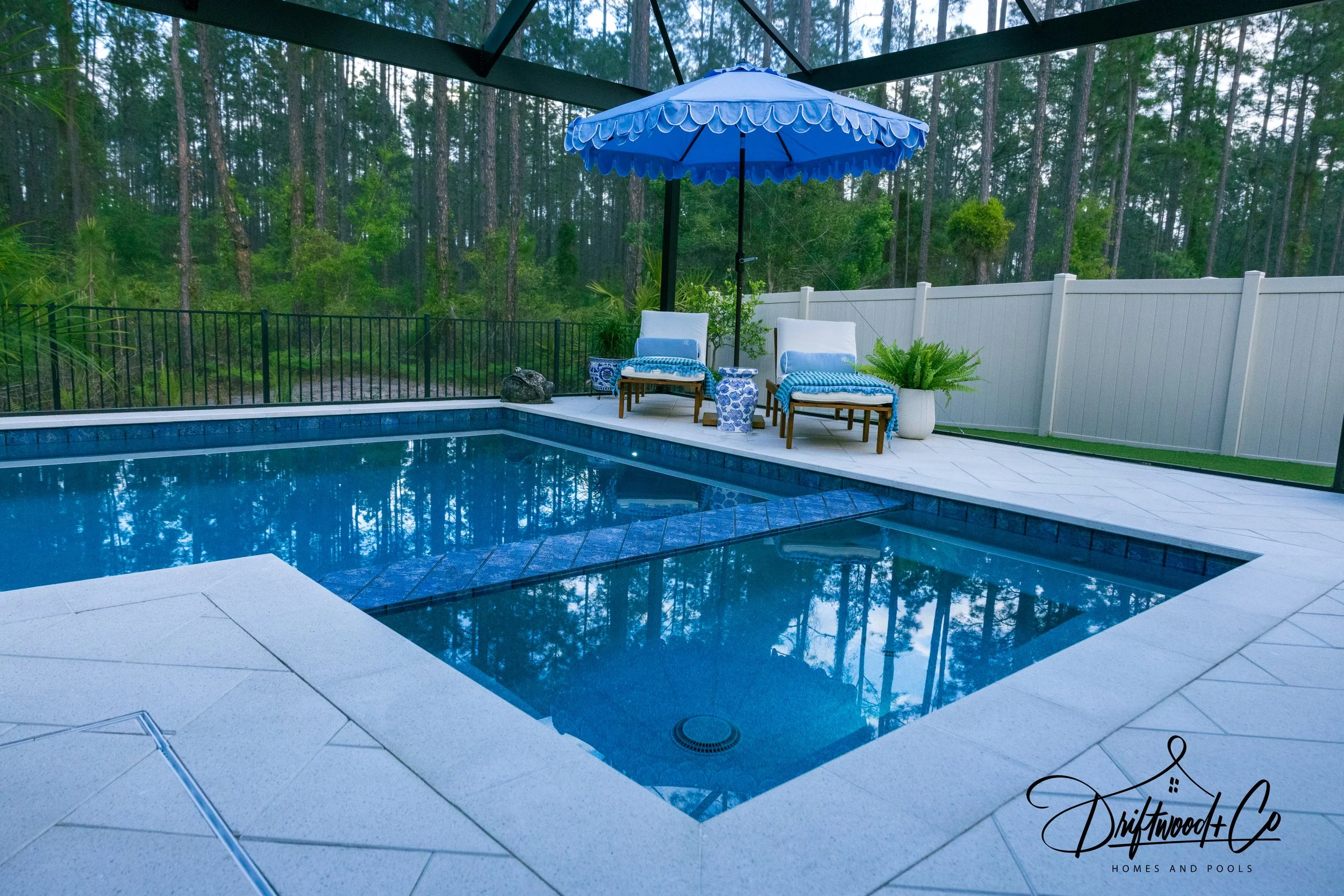 Swimming pool with two lounge chairs and a blue umbrella, surrounded by a white fence and trees in the background.