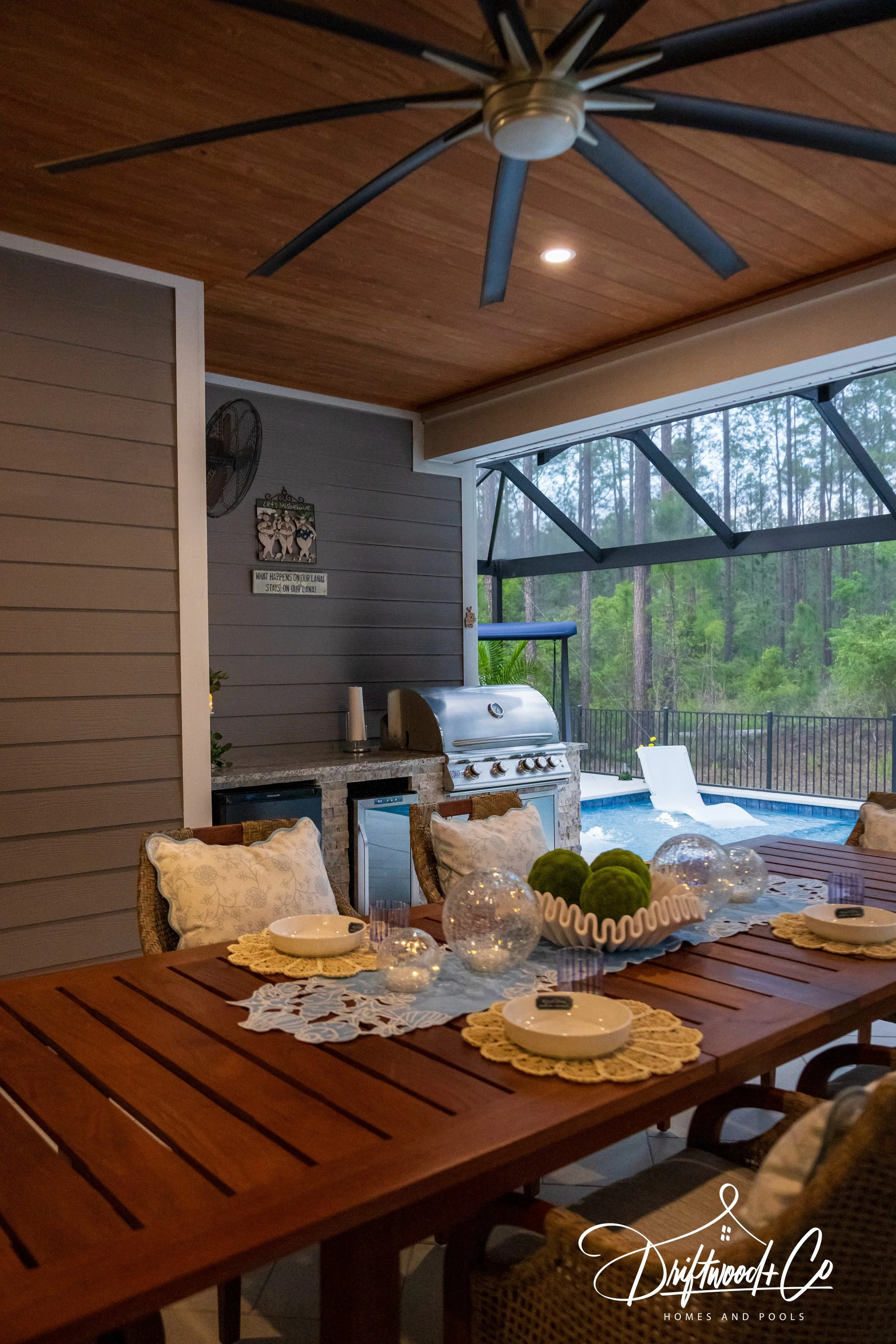 Outdoor patio with wooden dining table set for a meal, decorated with a lace runner and centerpieces. Adjacent to a pool with a float, partially covered by a screened porch. Ceiling fan and wall-mounted fan in the background, with forest visible beyond. Interior and exterior space blending for outdoor living.