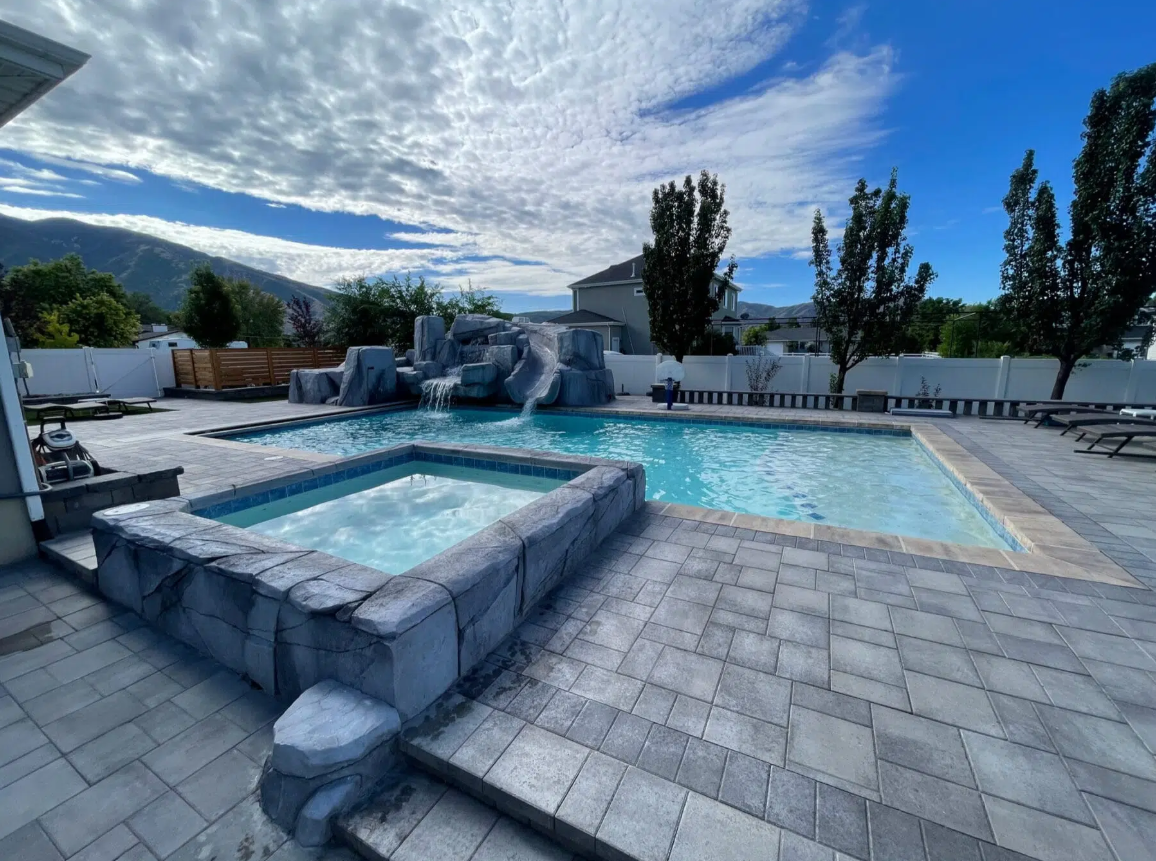 A backyard swimming pool with a rock waterfall feature, surrounded by a paved deck with lounge chairs, trees, and a white fence. Mountainous landscape and blue sky with clouds in the background.