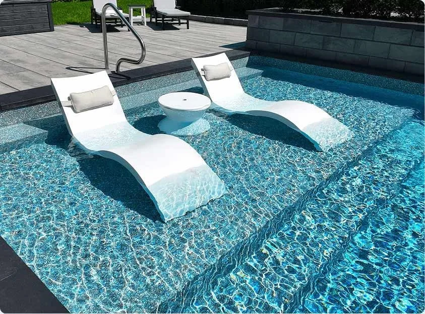 Two white pool lounge chairs with pillows and a small round table in a swimming pool.