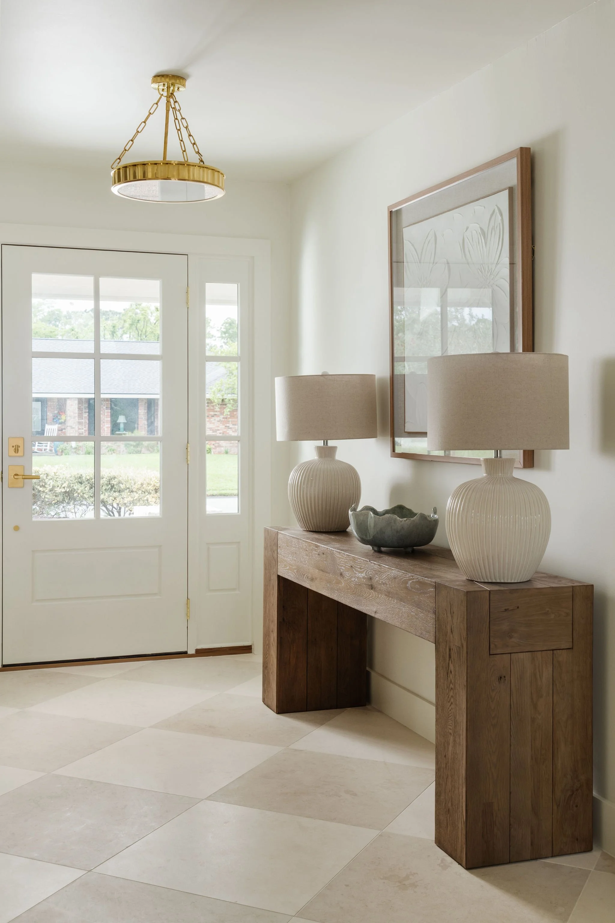 Entryway with white door, wooden console table topped with two beige lamps and decorative bowl, framed artwork on the wall, and a brass chandelier hanging from the ceiling.