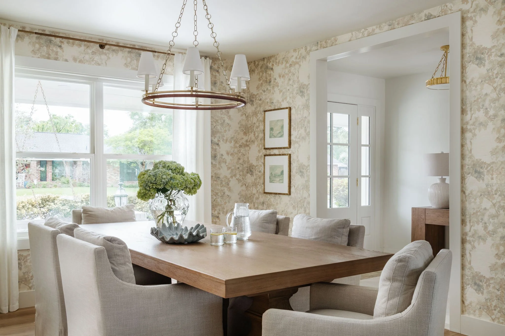 A dining room with a large wooden table surrounded by upholstered chairs, a vase of green hydrangeas, large windows with white curtains, floral-patterned wallpaper, framed artwork, and a circular chandelier with interior lights.