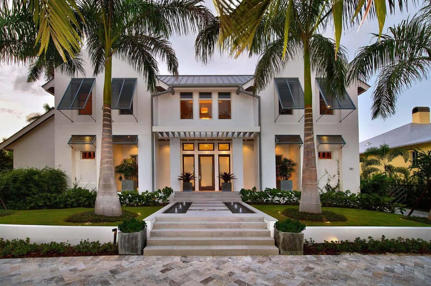 Front view of a modern white house with a staircase leading to the entrance, surrounded by lush green landscaping, two tall palm trees flanking the walkway, and large windows with black shutters.