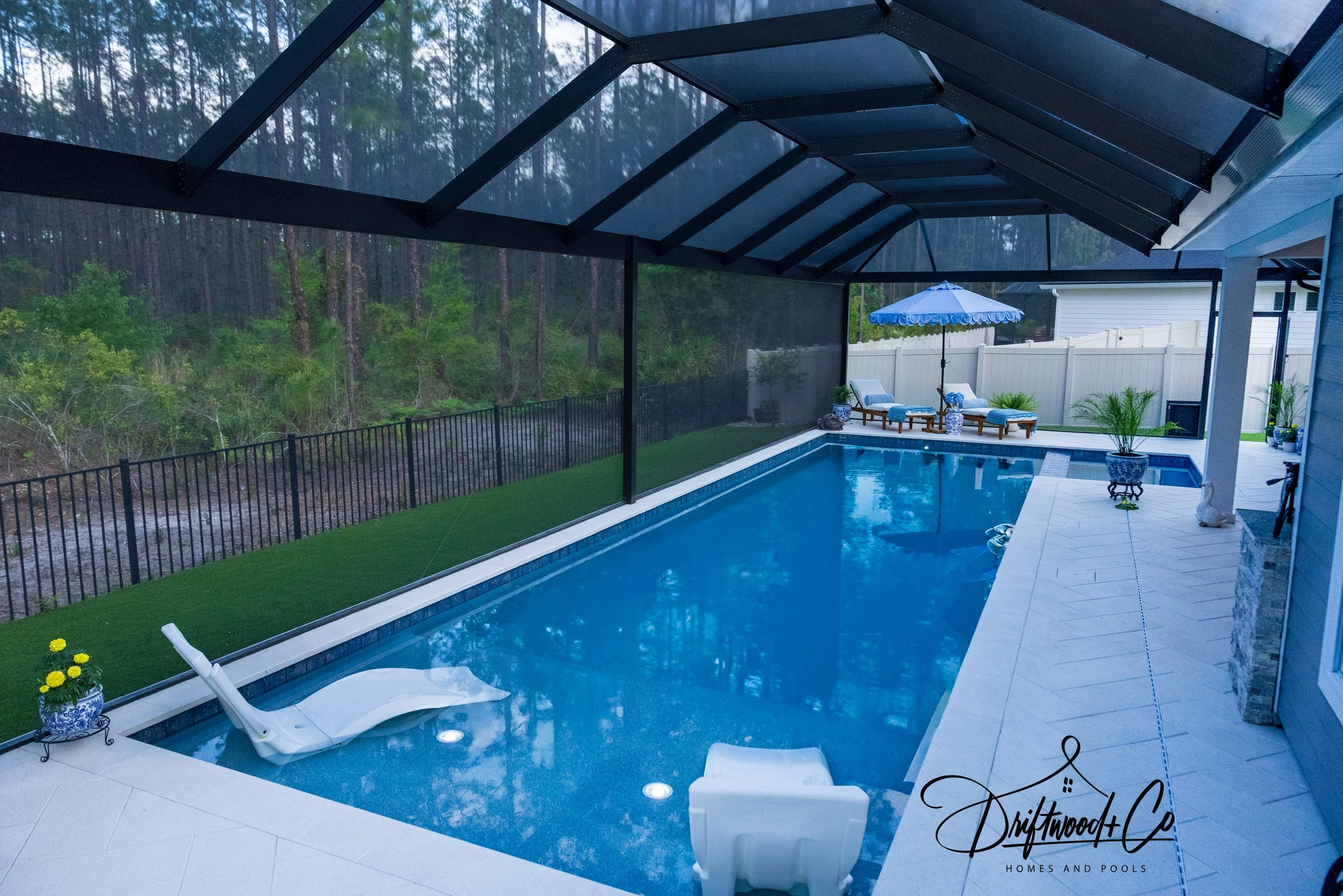 A backyard swimming pool with a white lounge chair in the foreground, decorated with potted plants and a glow-in-the-dark sign that reads 'Driftwood & Co. Homes and Pools.' in the corner. There are seating areas with umbrellas and wooden lounge chairs along the poolside, and a wooded area with trees and greenery is visible behind a black safety fence.