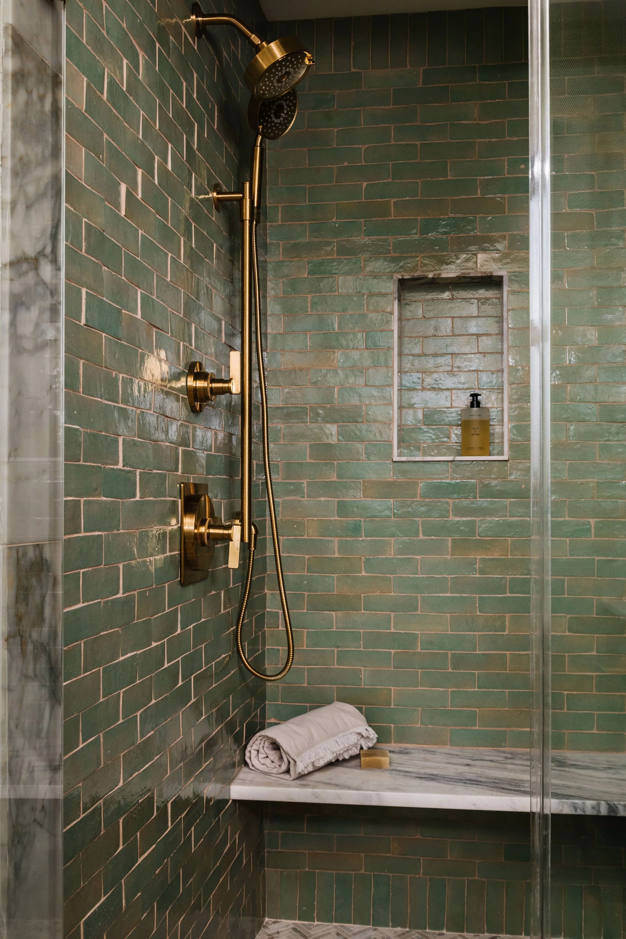 A modern shower with green subway tile walls, a gold showerhead and fixtures, a built-in niche with a soap dispenser, a folded towel and a bar of soap on a marble shelf, and a marble floor.
