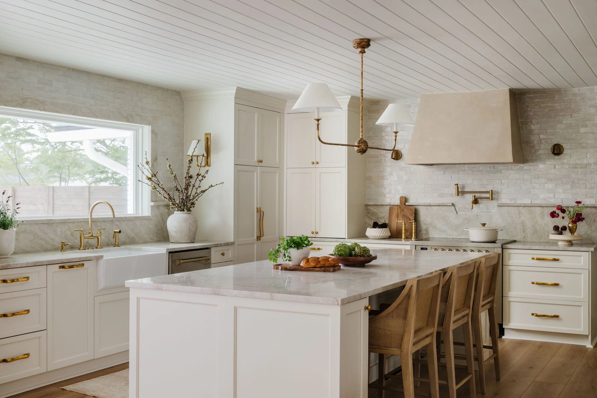 Modern kitchen with white cabinets, marble countertops, gold hardware, a large window, and a chandelier with five lampshades hanging from the ceiling.