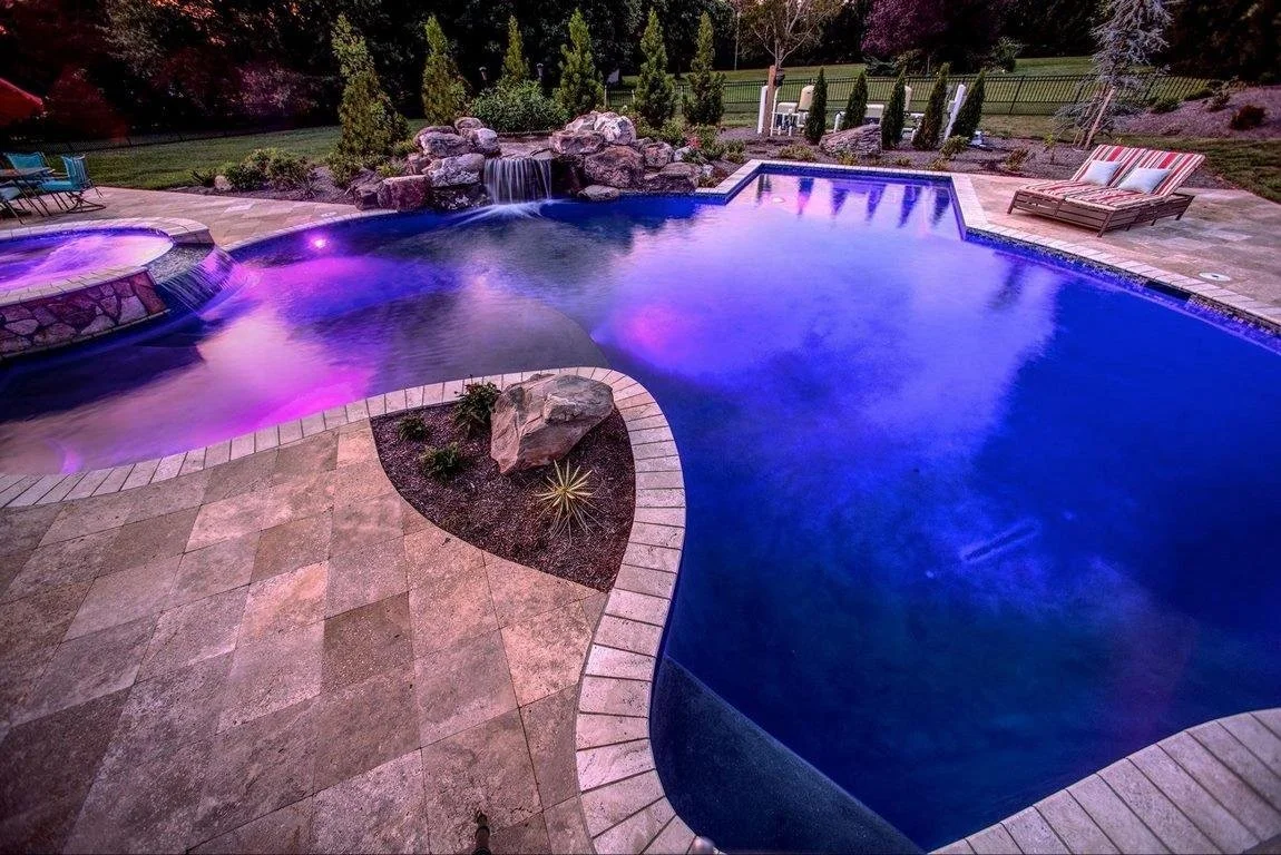 Nighttime view of a backyard swimming pool with purple lights and a hot tub, surrounded by stone patio, rocks, waterfall, trees, and outdoor furniture.