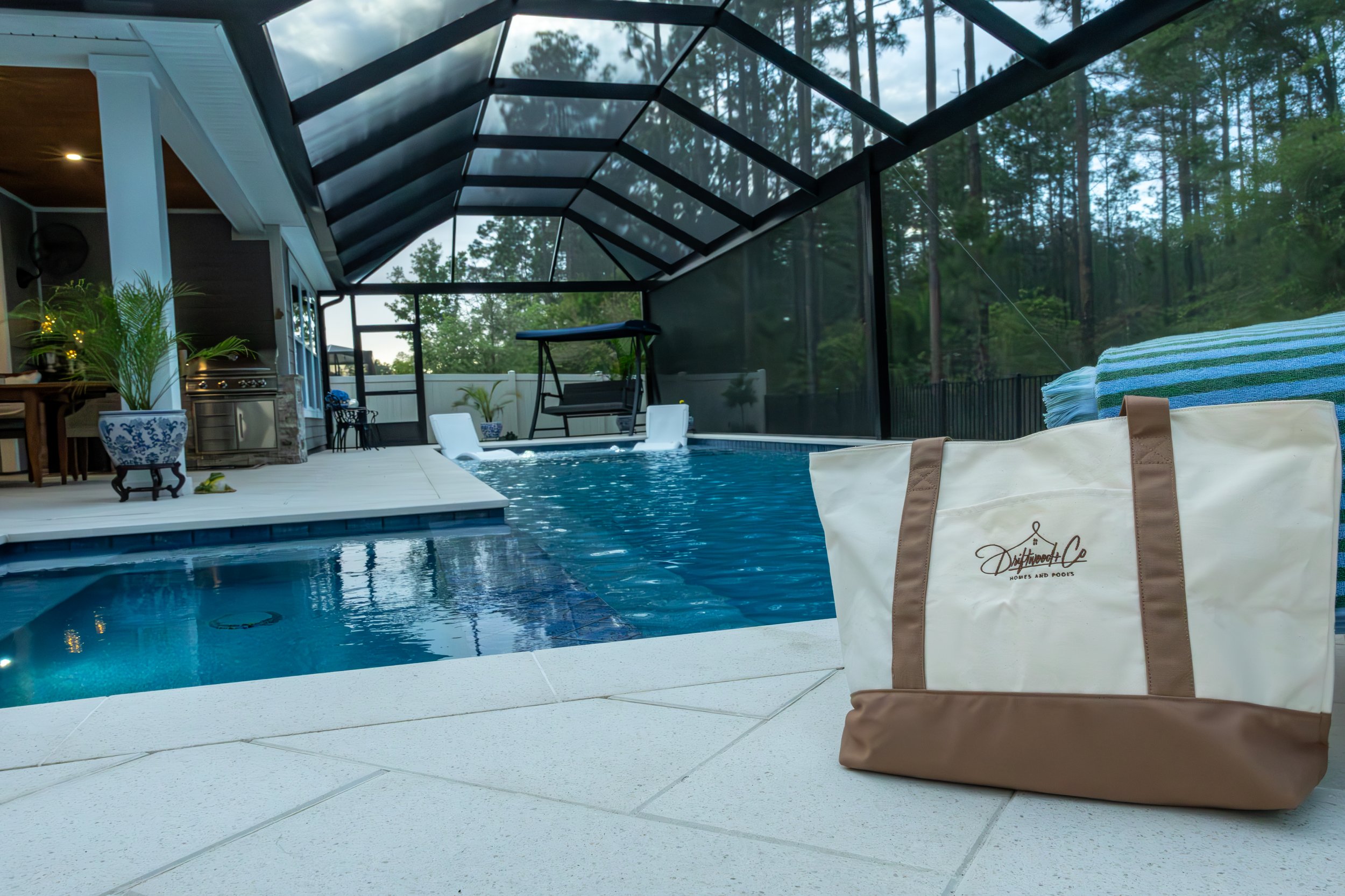 View of a screened-in outdoor pool area with a shaded enclosure, a swimming pool, a poolside lounge chair, a pao bag with the logo 'Westwood & Co Homes and Pools', and surrounding trees.