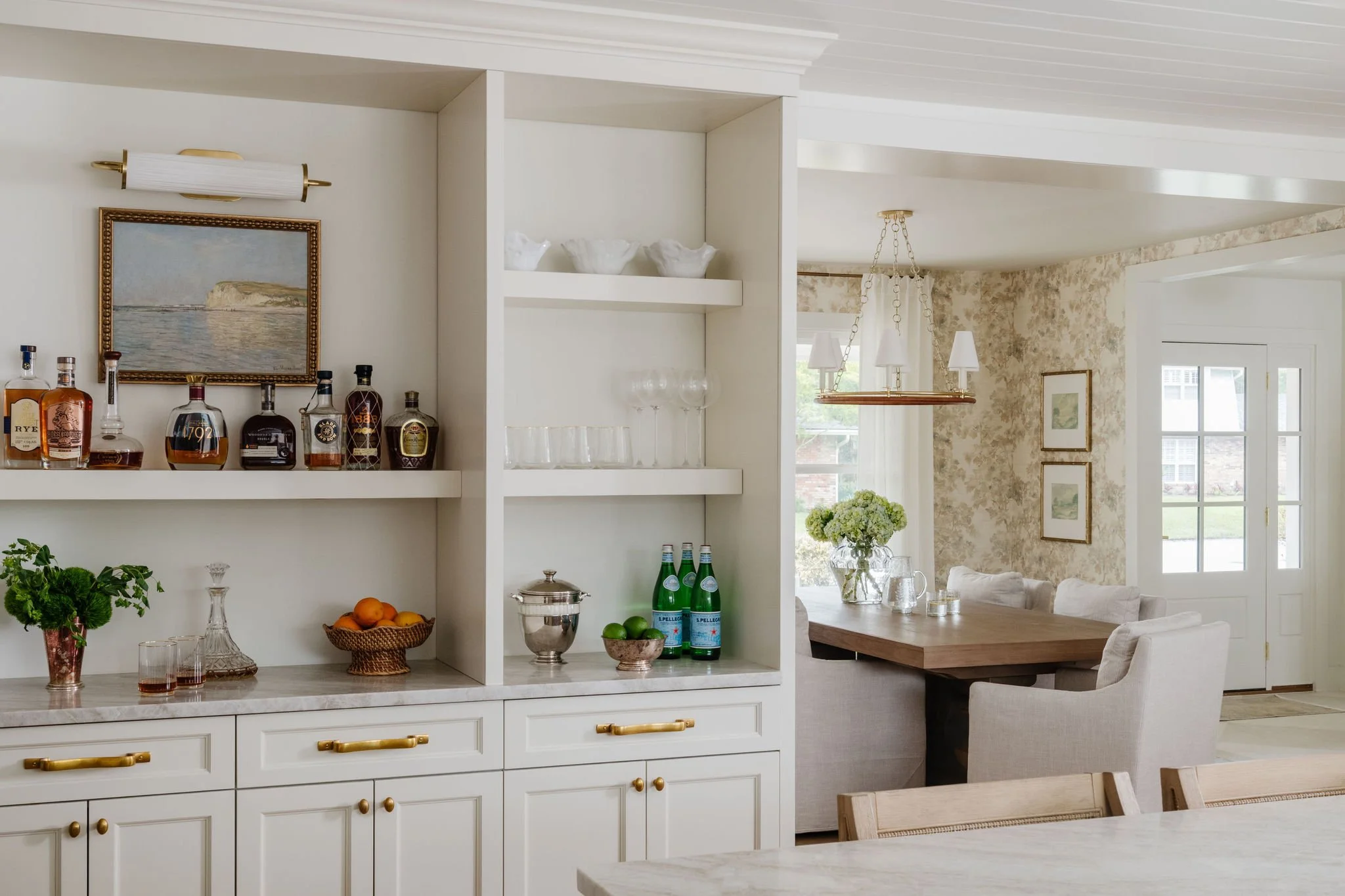 Kitchen and dining area with white cabinetry, a bookshelf with bottles of alcohol, glassware, fruit, and beverages, and a bright, airy living room with large windows, framed artwork, and a chandelier.
