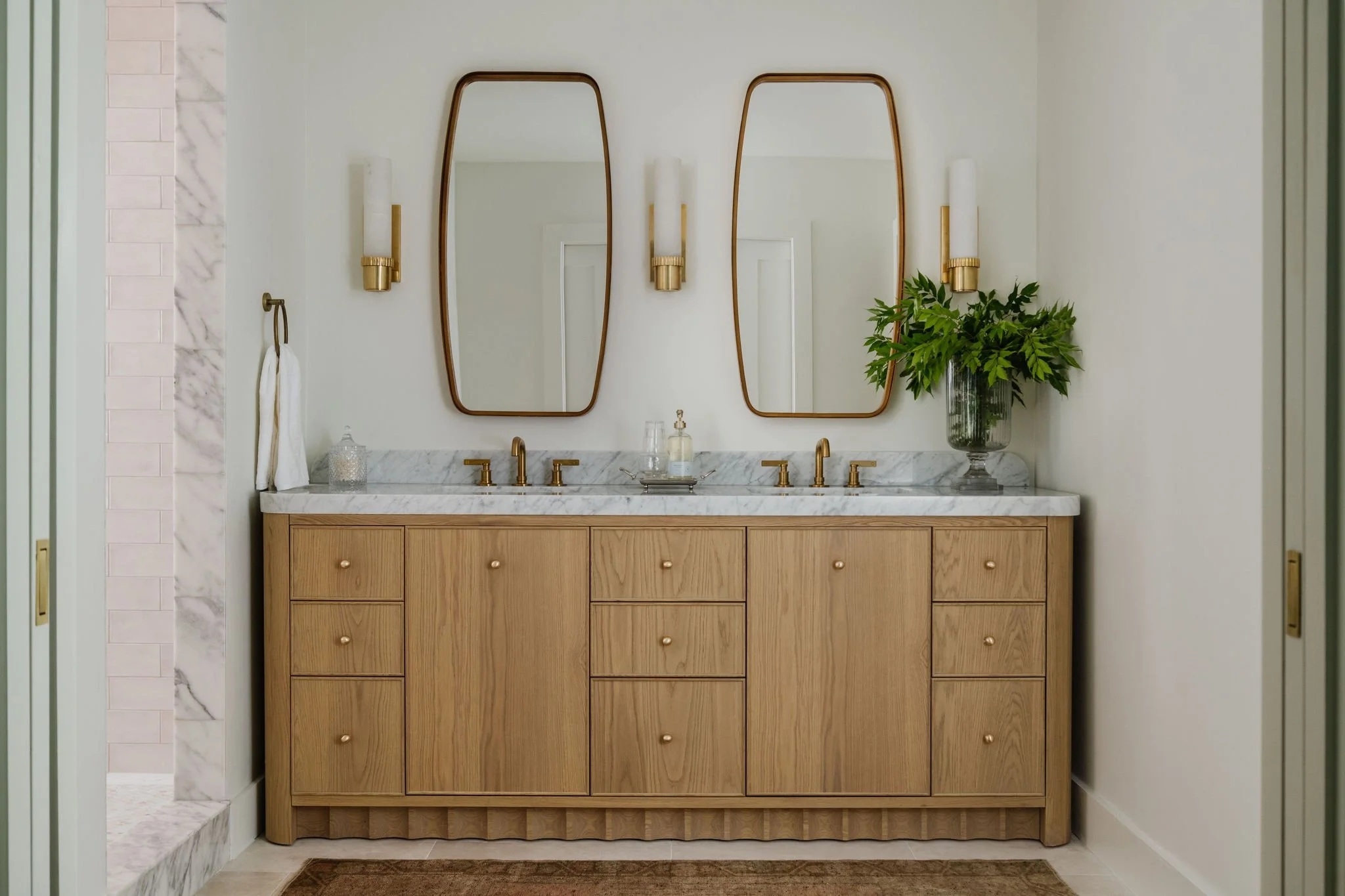 Bathroom vanity with a marble countertop, two gold faucets, a large green plant in a glass vase, and two mirrors with gold frames, with wall-mounted gold and white light fixtures on either side.