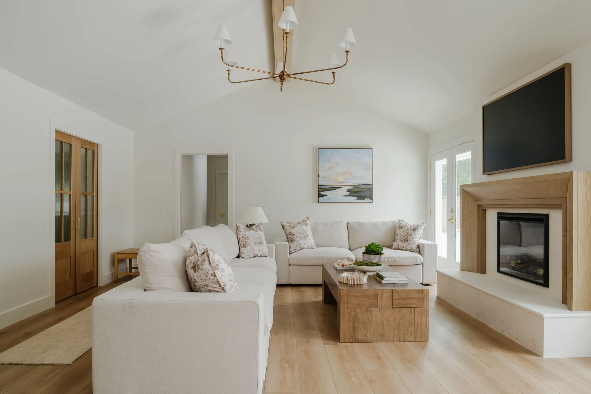 Bright living room with white walls, a light-colored sofa and armchair with floral pillows, a wooden coffee table, a fireplace with a wooden surround, a wall-mounted TV, artwork, and a modern chandelier.