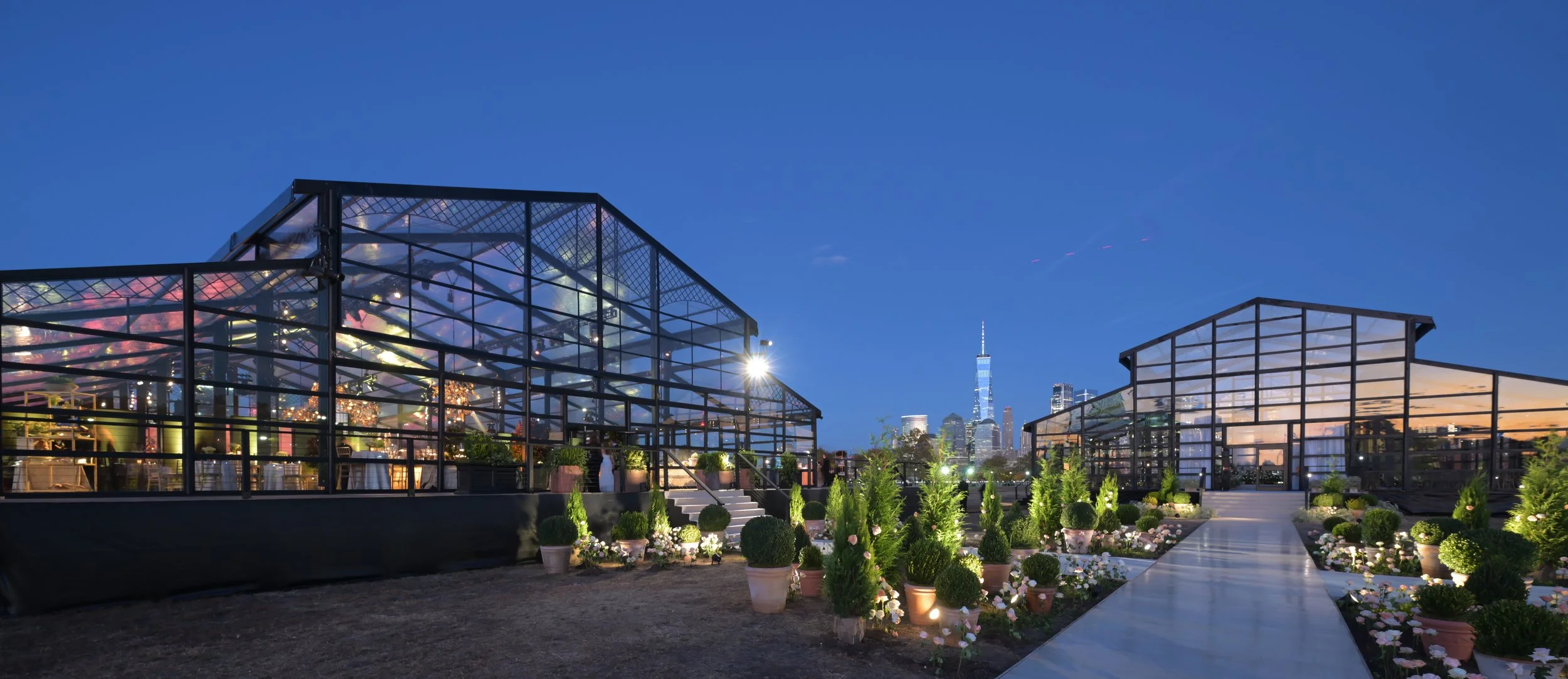 Nighttime view of two glass greenhouses on a rooftop garden, with Manhattan skyline including the One World Trade Center in the background. The garden has potted plants and decorative flowers along a walkway.